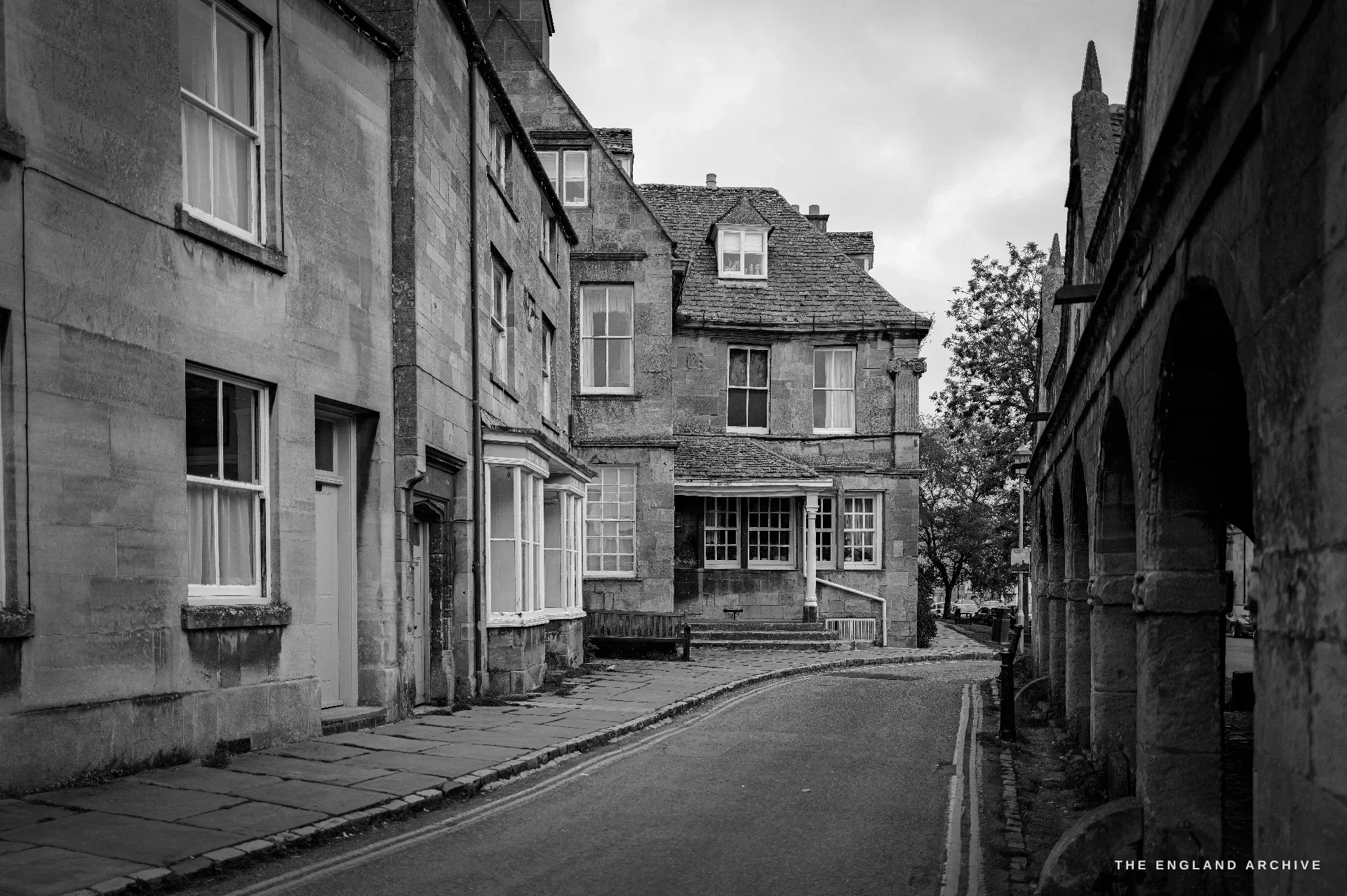 A quiet sloping back-street of terraced Cotswold-stone houses with sash bay windows, the road curving away past the edge of the Market Hall arcade.