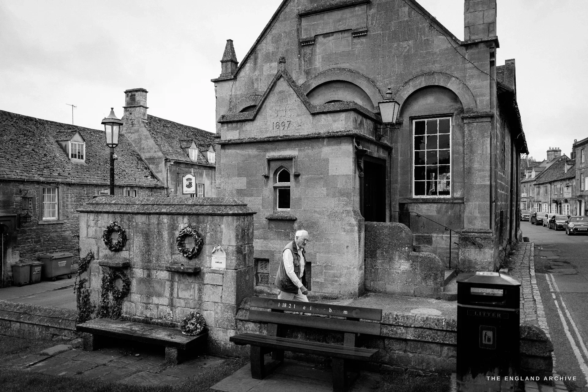 A dated stone almshouse with arched windows and gables, an elderly man pausing on a bench beside framed wreaths on a low wall, the date 1897 carved in the gable.