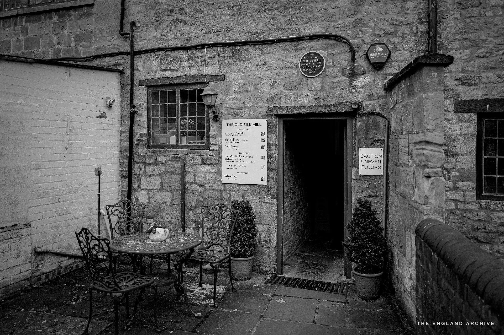 The cobbled courtyard entrance to the Old Silk Mill, a stone doorway under a round blue plaque, a hand-lettered notice board and a cafe table set with a teapot.
