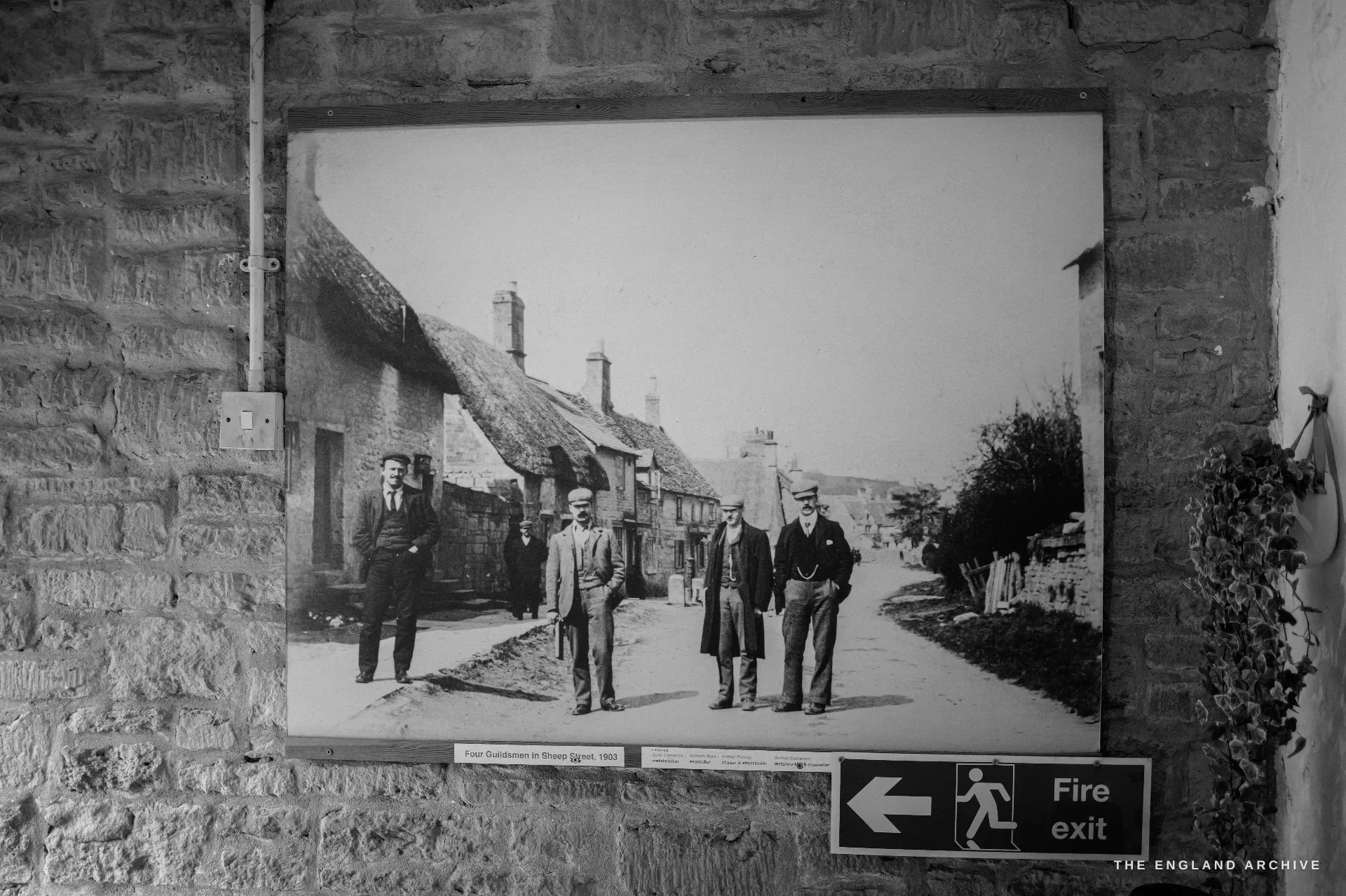 A large mounted archival photograph on the Mill wall showing Edwardian Guild craftsmen standing in a Campden street, a green fire-exit sign below it.