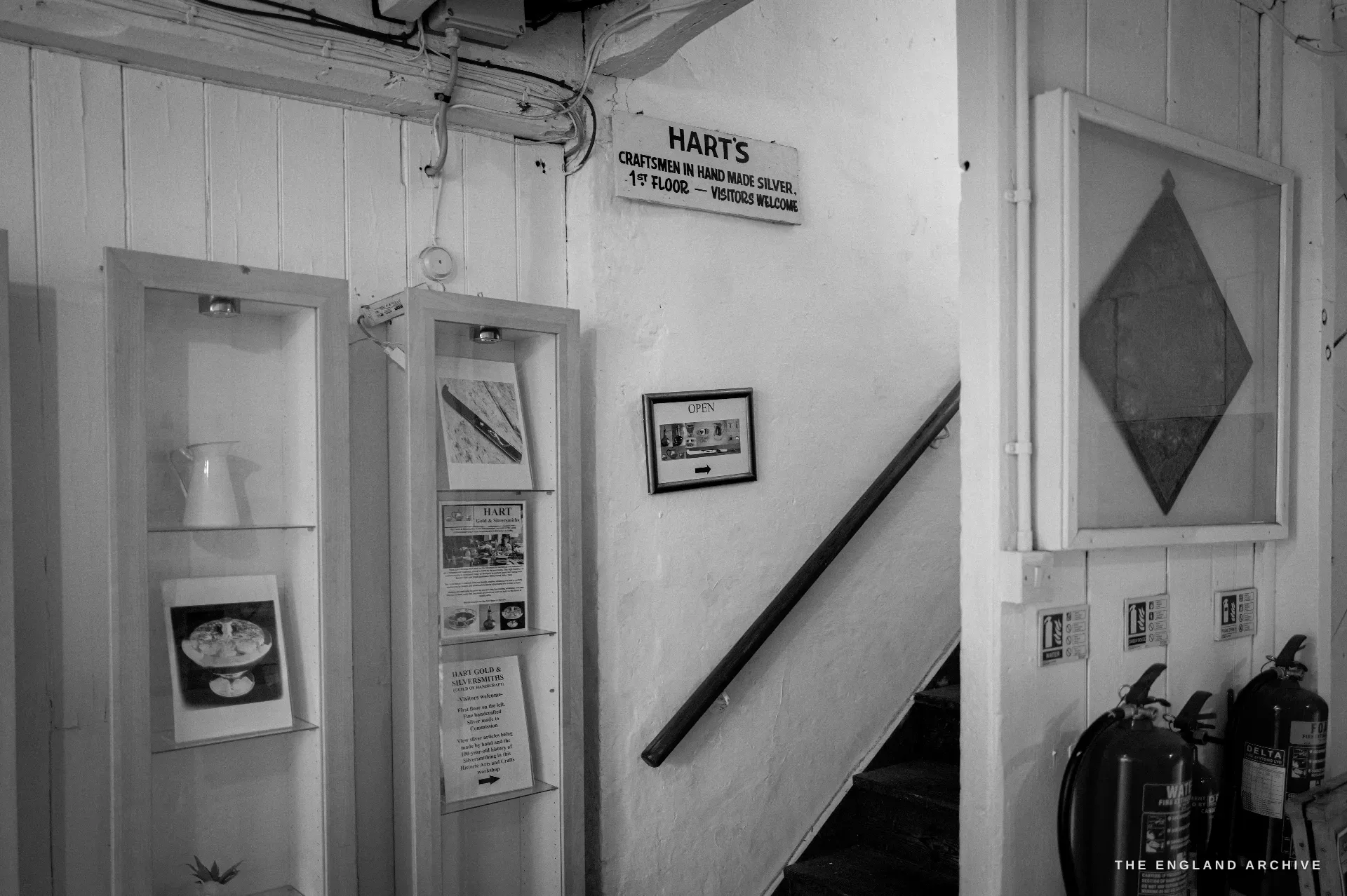 The workshop stairwell, display niches of silver and framed drawings on the white wall, beneath a hand-lettered sign reading Harts, Craftsmen in Hand Made Silver, Visitors Welcome.