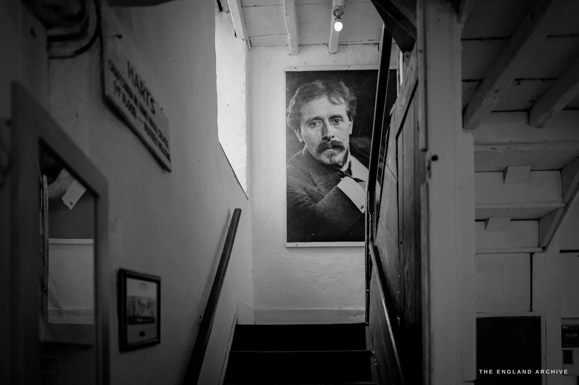 Looking up the worn workshop staircase to a large mounted portrait photograph of a bearded Edwardian man, lit from a skylight, framed pictures down the side wall.