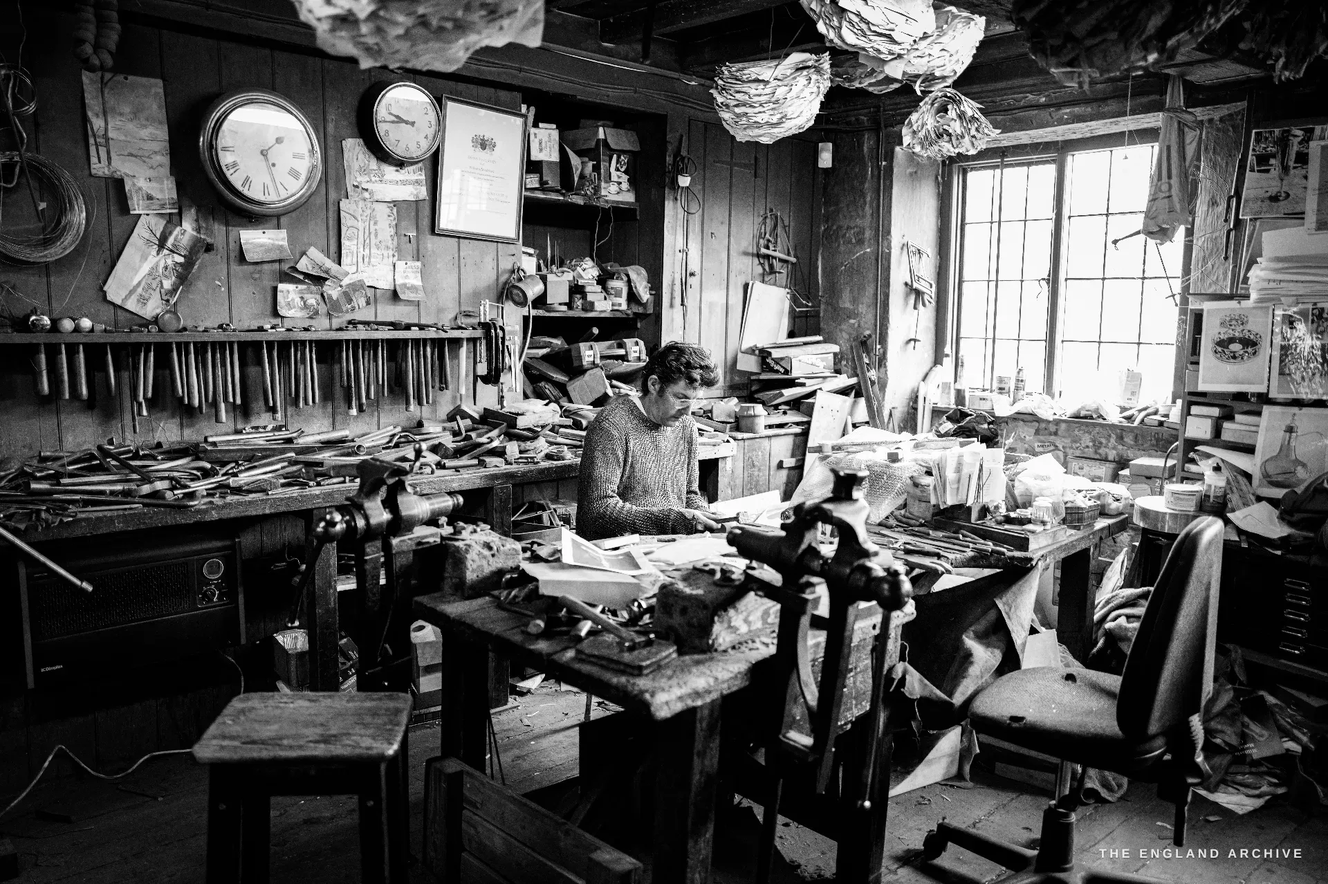 The wide, time-frozen bench room, a younger craftsman at the central bench amid racked hand tools, two wall clocks and a sunlit cottage window.