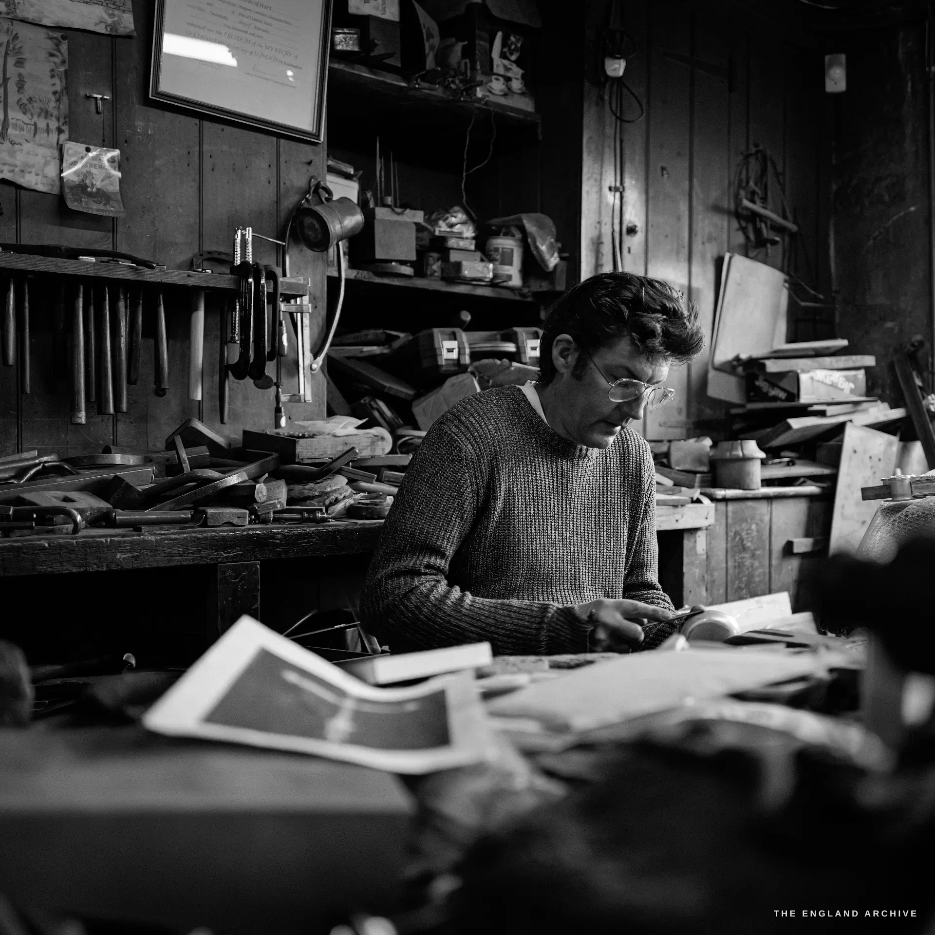 A square close portrait of a bespectacled younger craftsman in a chunky jumper bent over his bench, drawings spread before him, hand tools racked behind.