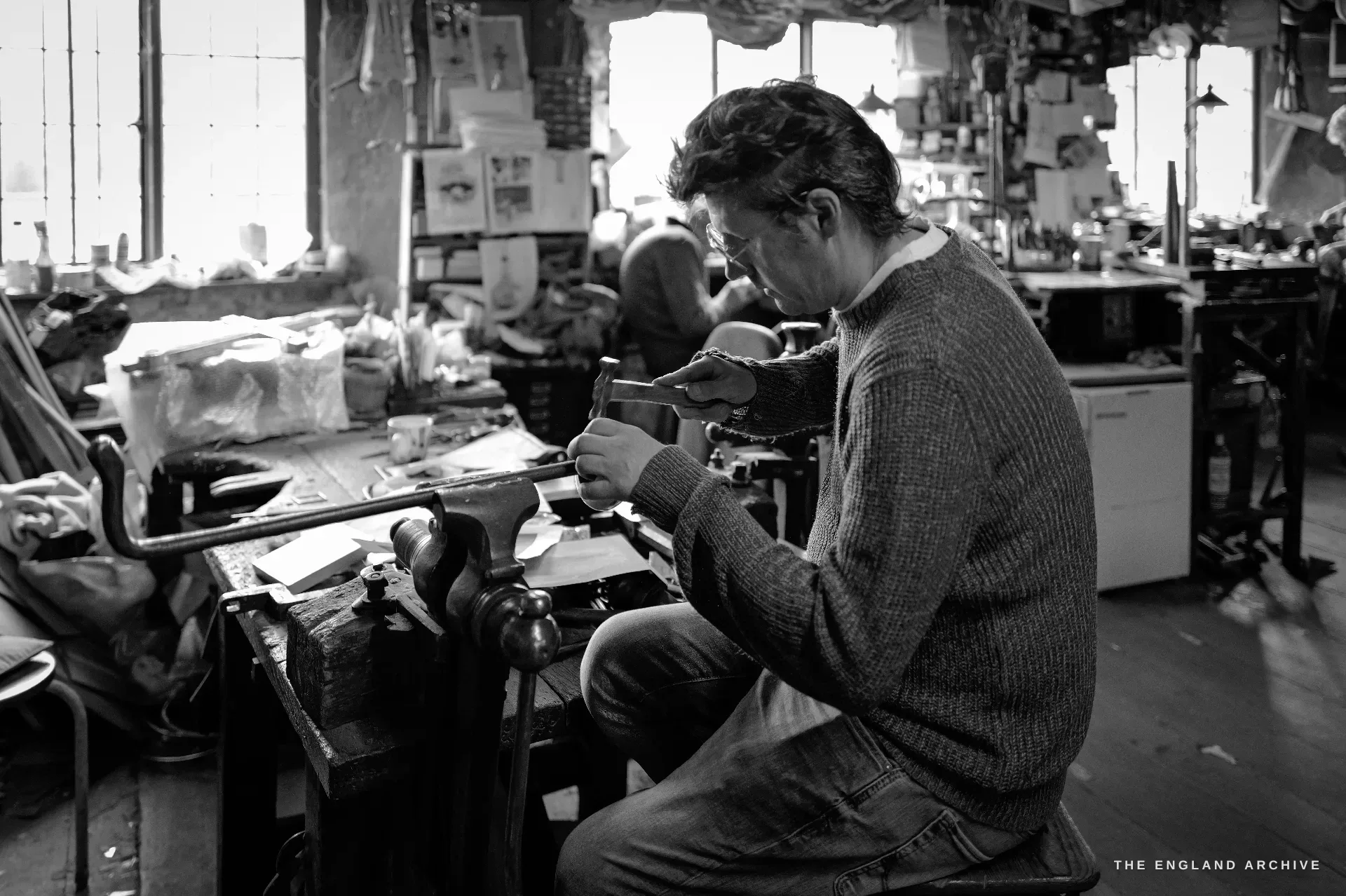 A younger craftsman sits at a jeweller's bench with its leather catch-skin and bench peg, holding a piece up to work it, a second worker dim in the sunlit background.