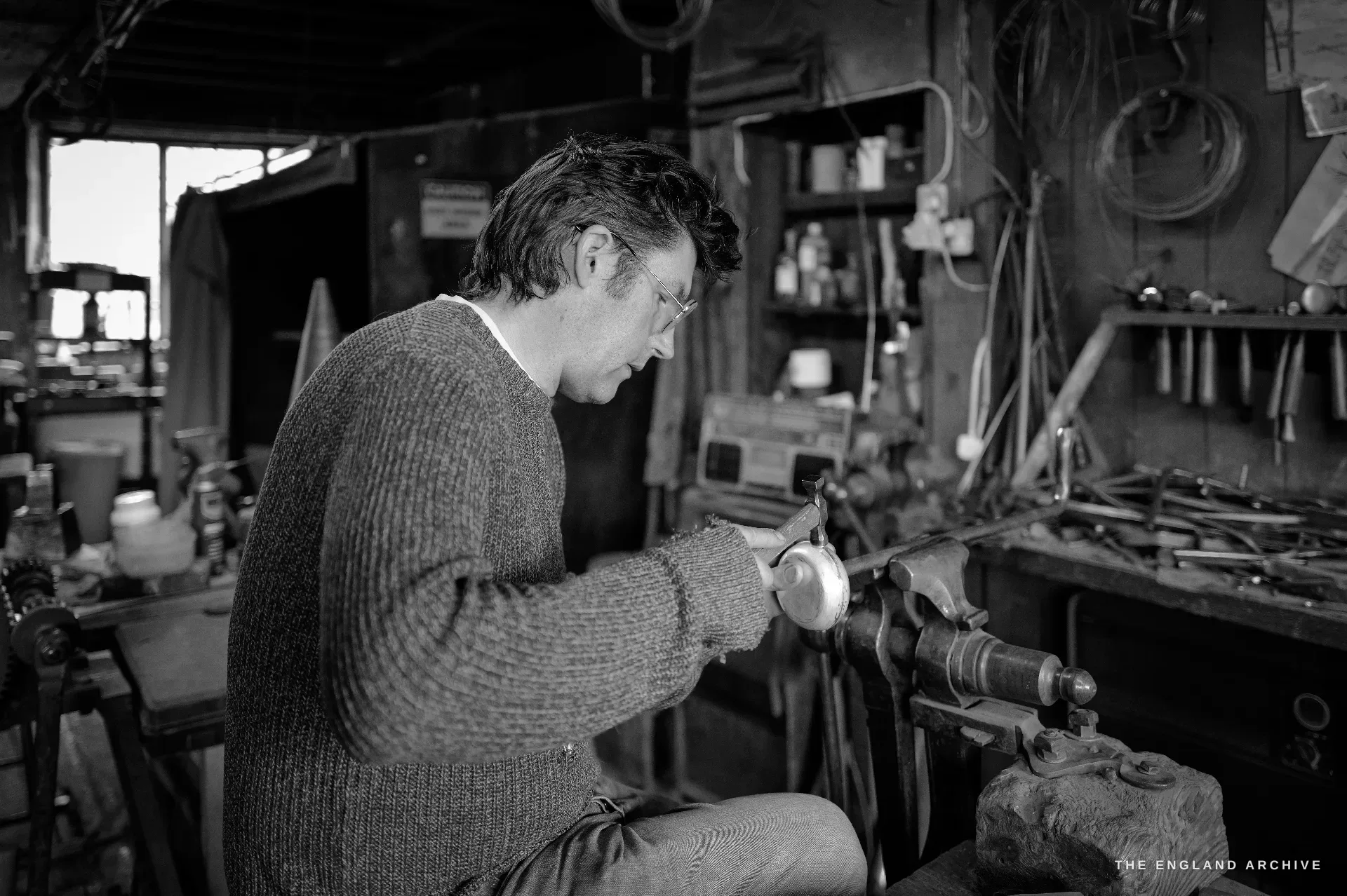A younger craftsman at a polishing spindle works a small silver bowl by hand, sleeves pushed up, the bench behind hung with pliers, files and coiled wire.