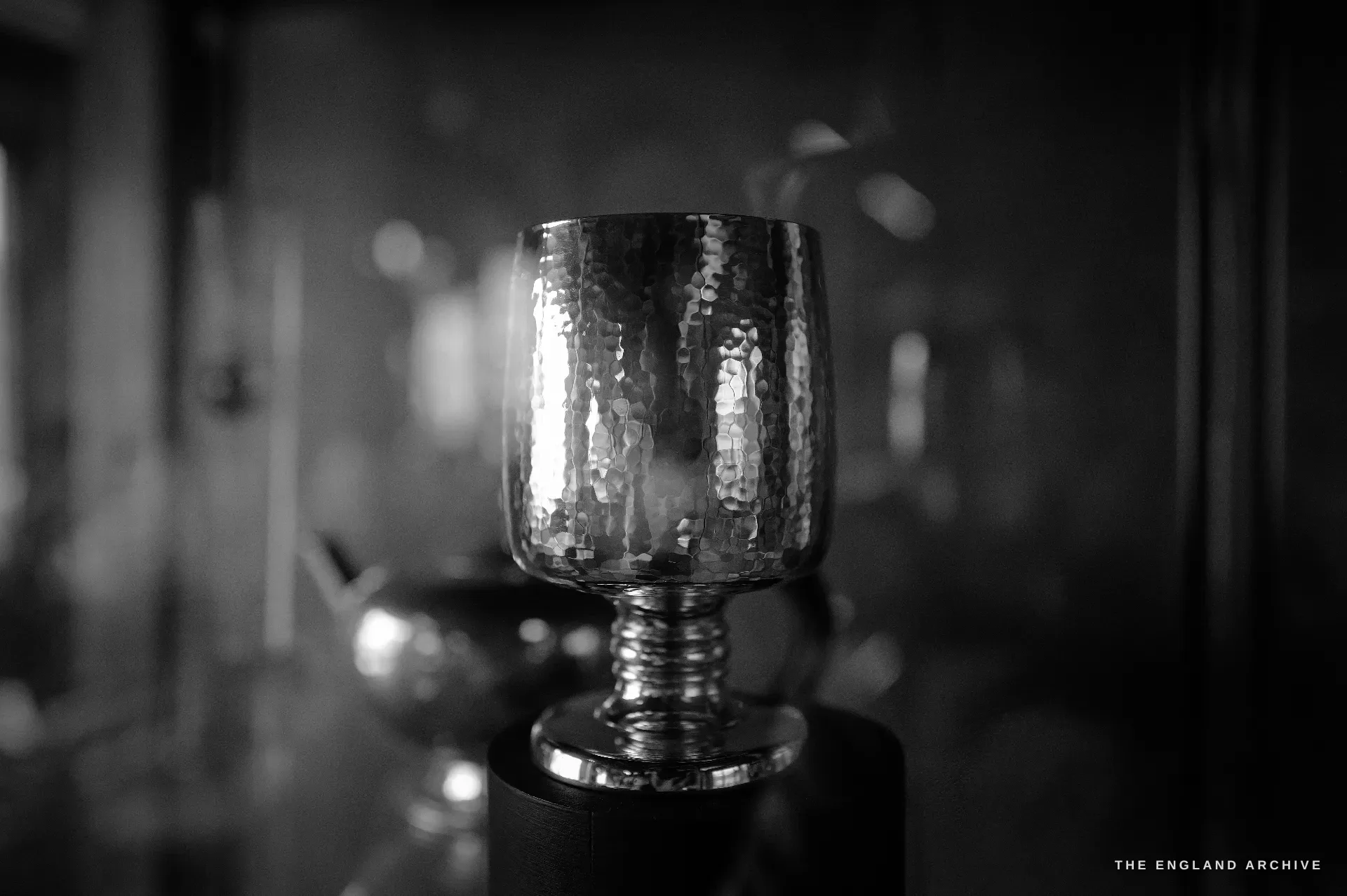 A single hammered-finish silver goblet on a baluster stem, lit on a black plinth, a second silver vessel softly out of focus behind it.