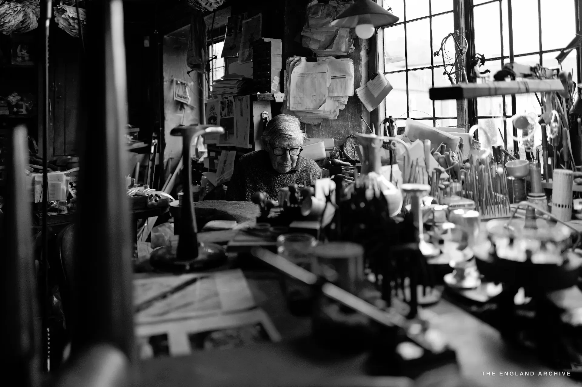 David Hart seated and bent over his bench beneath the big leaded window, surrounded by tools, jars and work-in-progress, framed through foreground clutter.