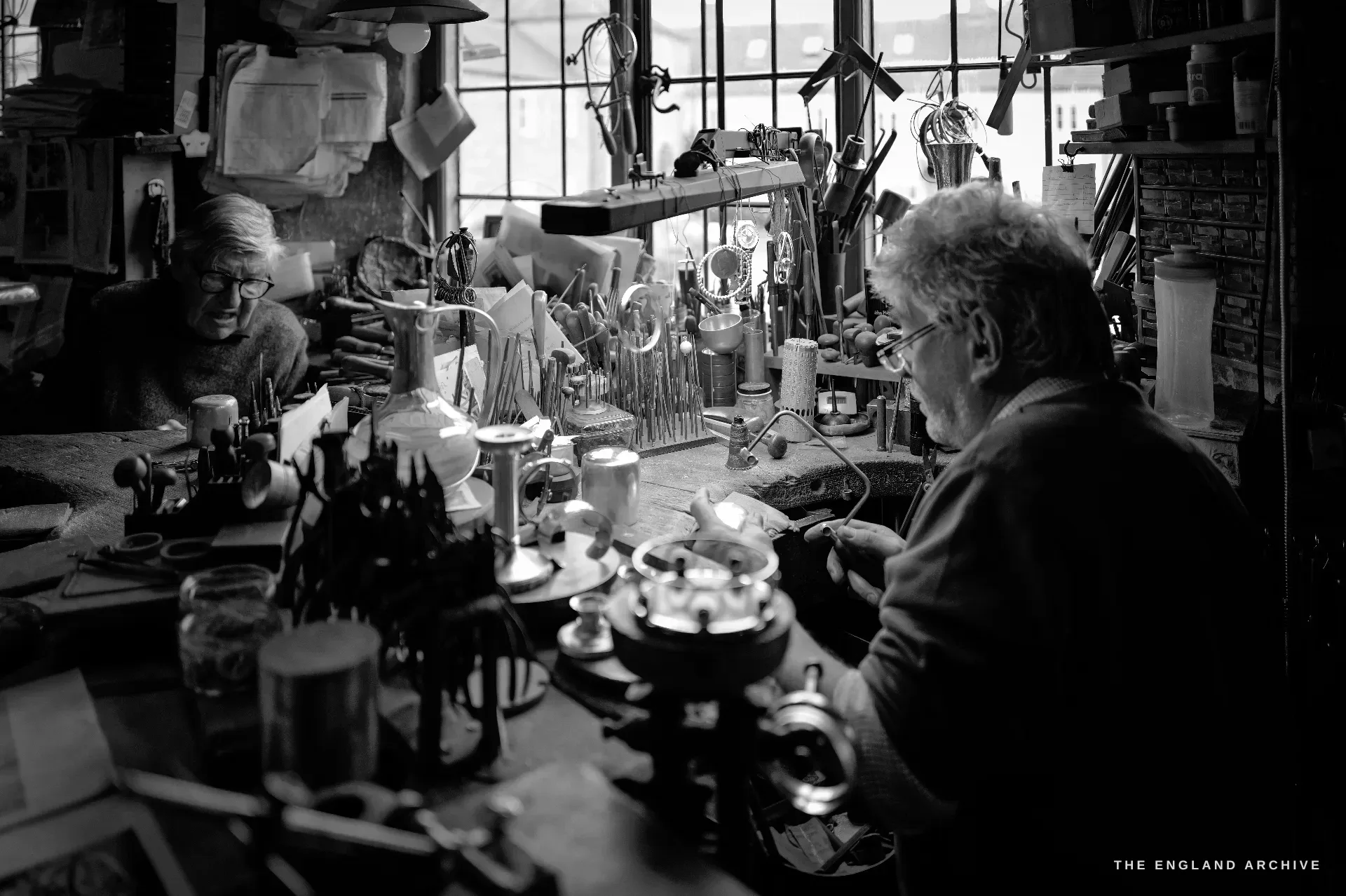 Two craftsmen at facing benches under the windows, the nearer man working under a bench lamp while the elder works at the far bench.