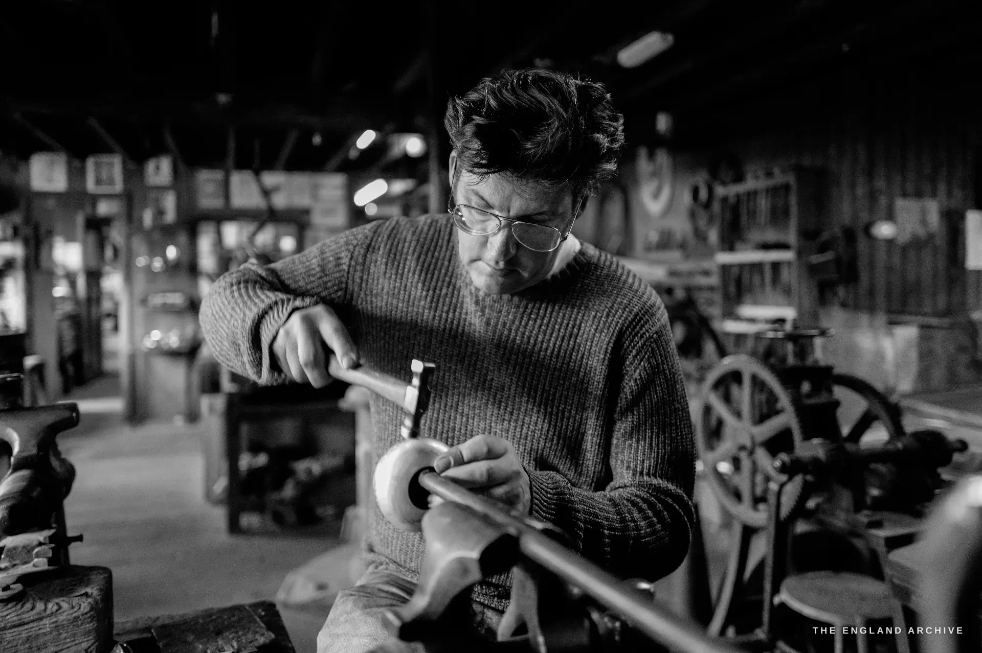 A younger craftsman in a wool jumper and round glasses works a piece on a spindle with a hammer, the workshop machinery and a large flywheel behind him.