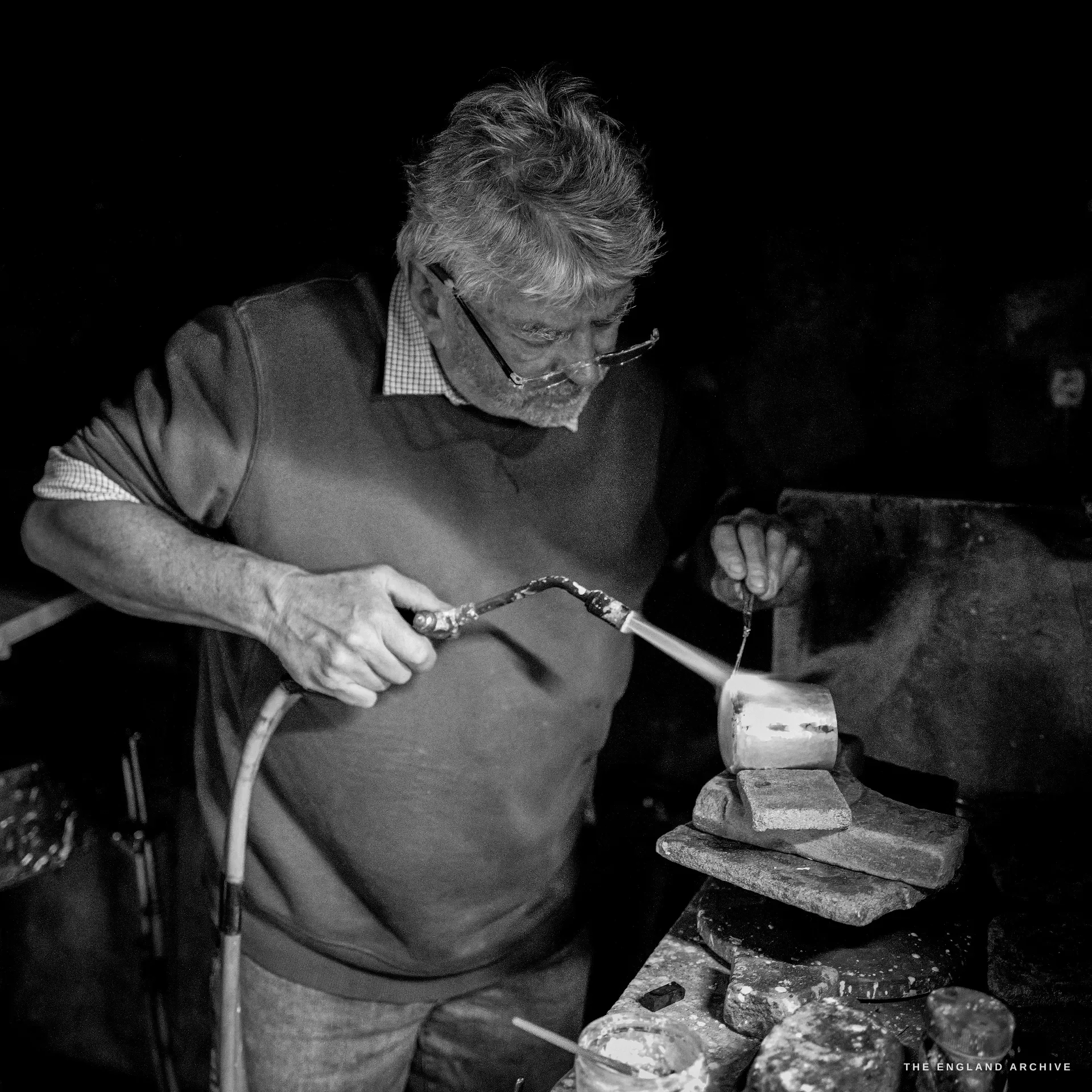A grey-haired craftsman in glasses handles metal with tongs at a casting station, ingots on the bench before him in the dark surround.