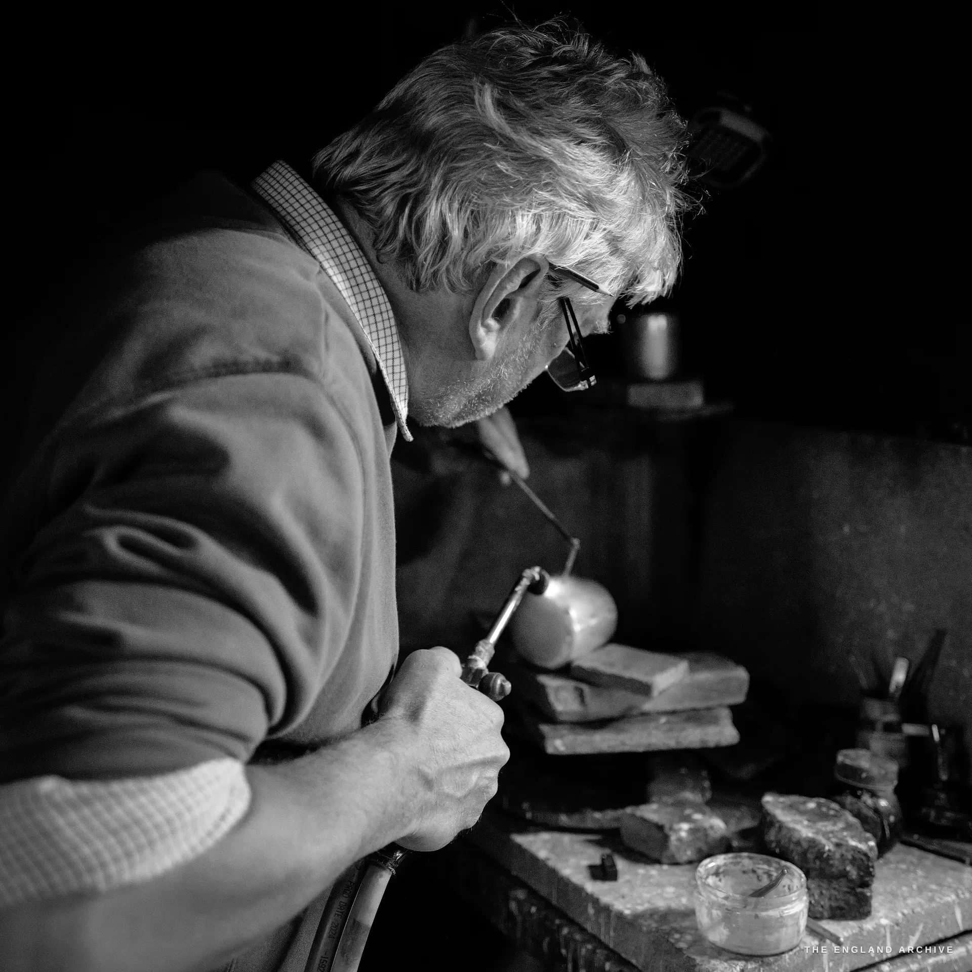 A tight side-profile of a grey-haired craftsman directing a small torch onto a rounded silver vessel held in his hand, files and blocks on the bench.