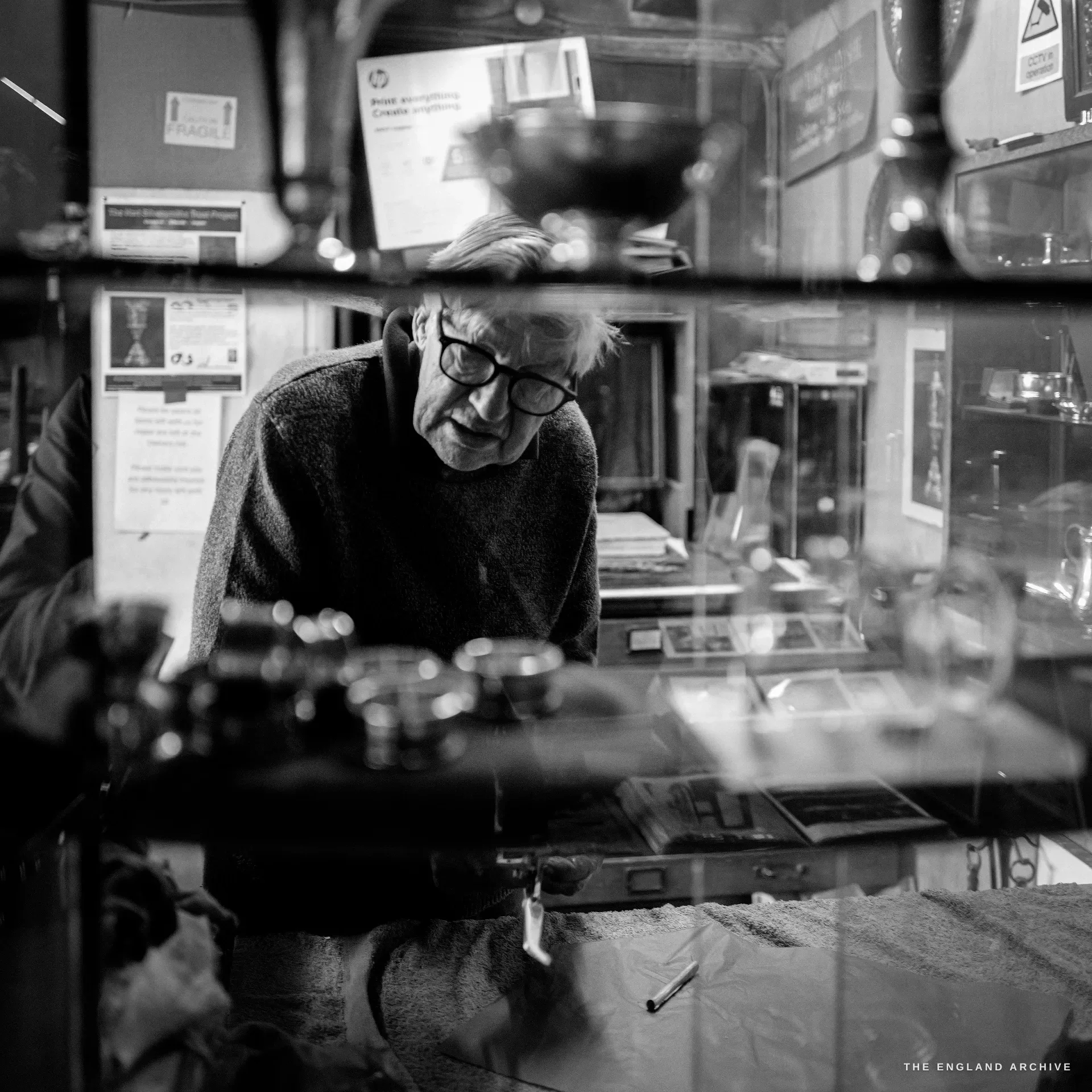 David Hart in heavy black-framed glasses leans over a glass display counter, seen through the reflective glass with a wineglass and silver pieces in the foreground.