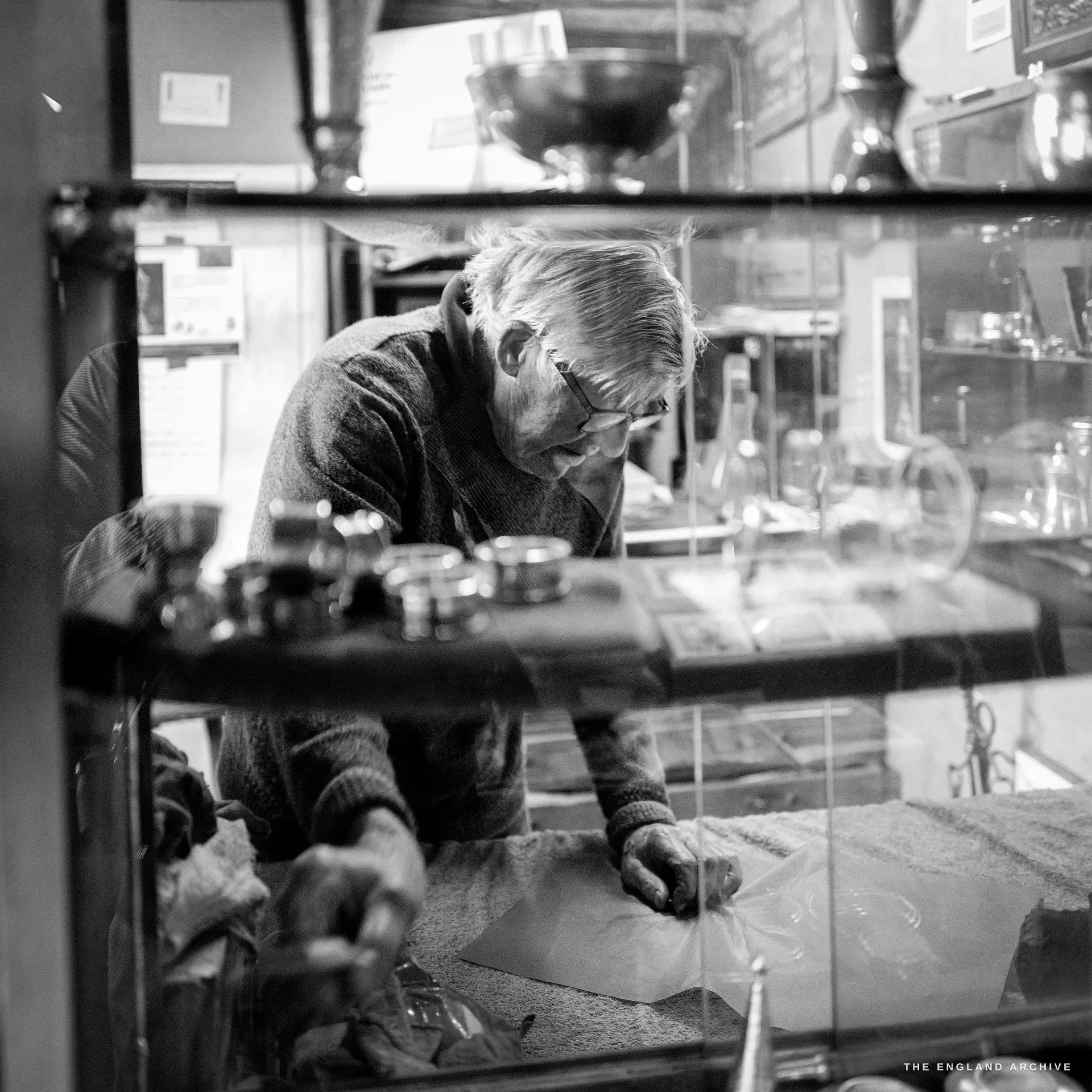 The elderly silversmith bent over the display case, smoothing tissue over silver salts with both hands, footed bowls on the shelf above, shot through the showcase glass.