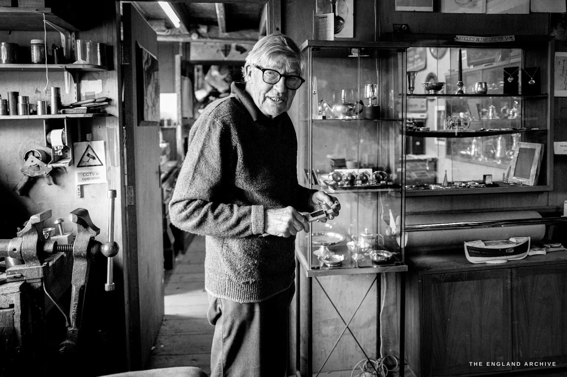 A full-length portrait of David Hart standing in the workshop in a grey jumper, a hand resting near a glazed cabinet of silver, a bench vice and tools at the left.
