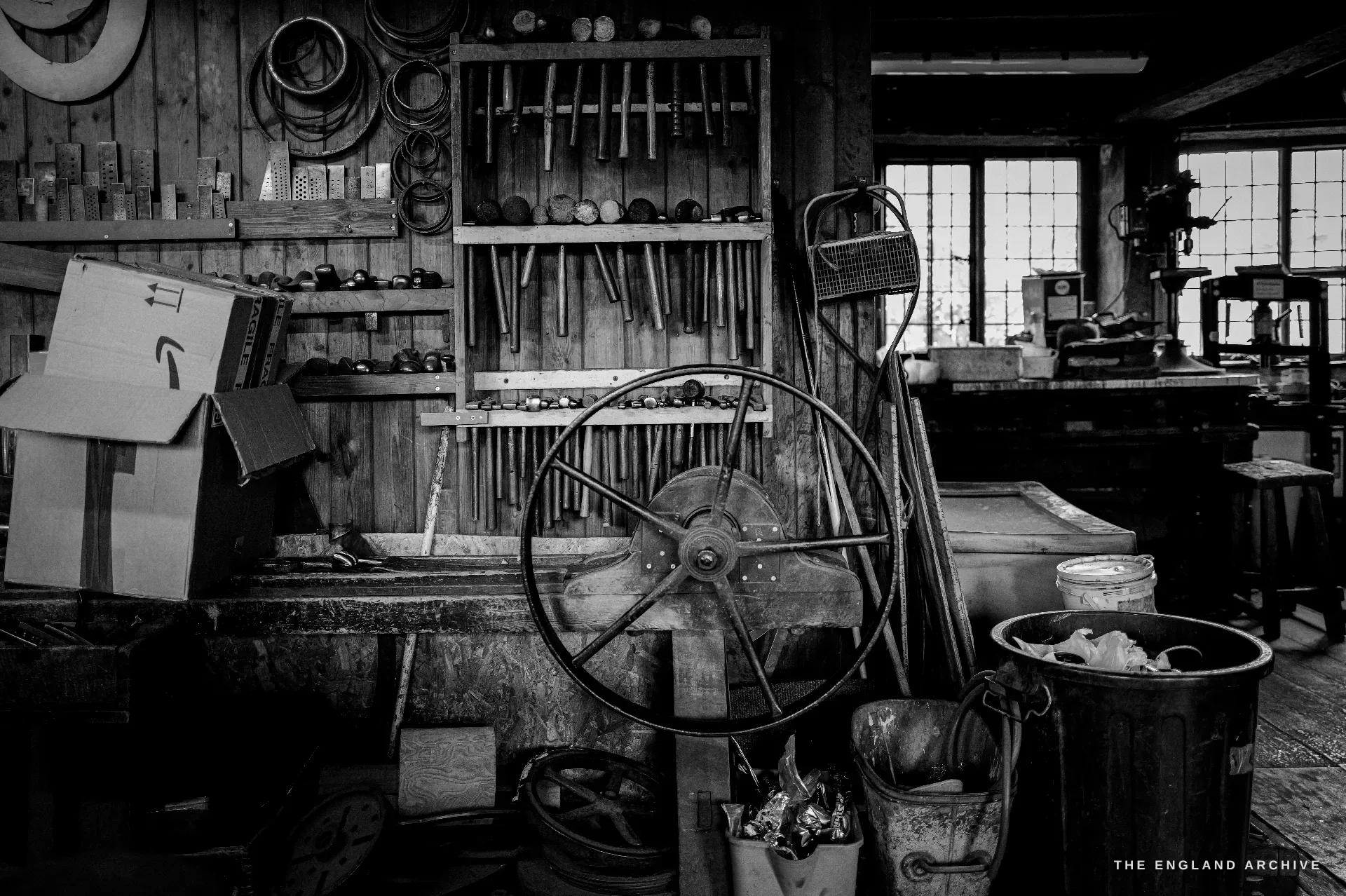 A frozen-in-time bench room: a large hand-cranked flywheel centre-frame, a board of files and stakes, hoops and patterns hung above, a galvanised bin and buckets, the leaded window behind.