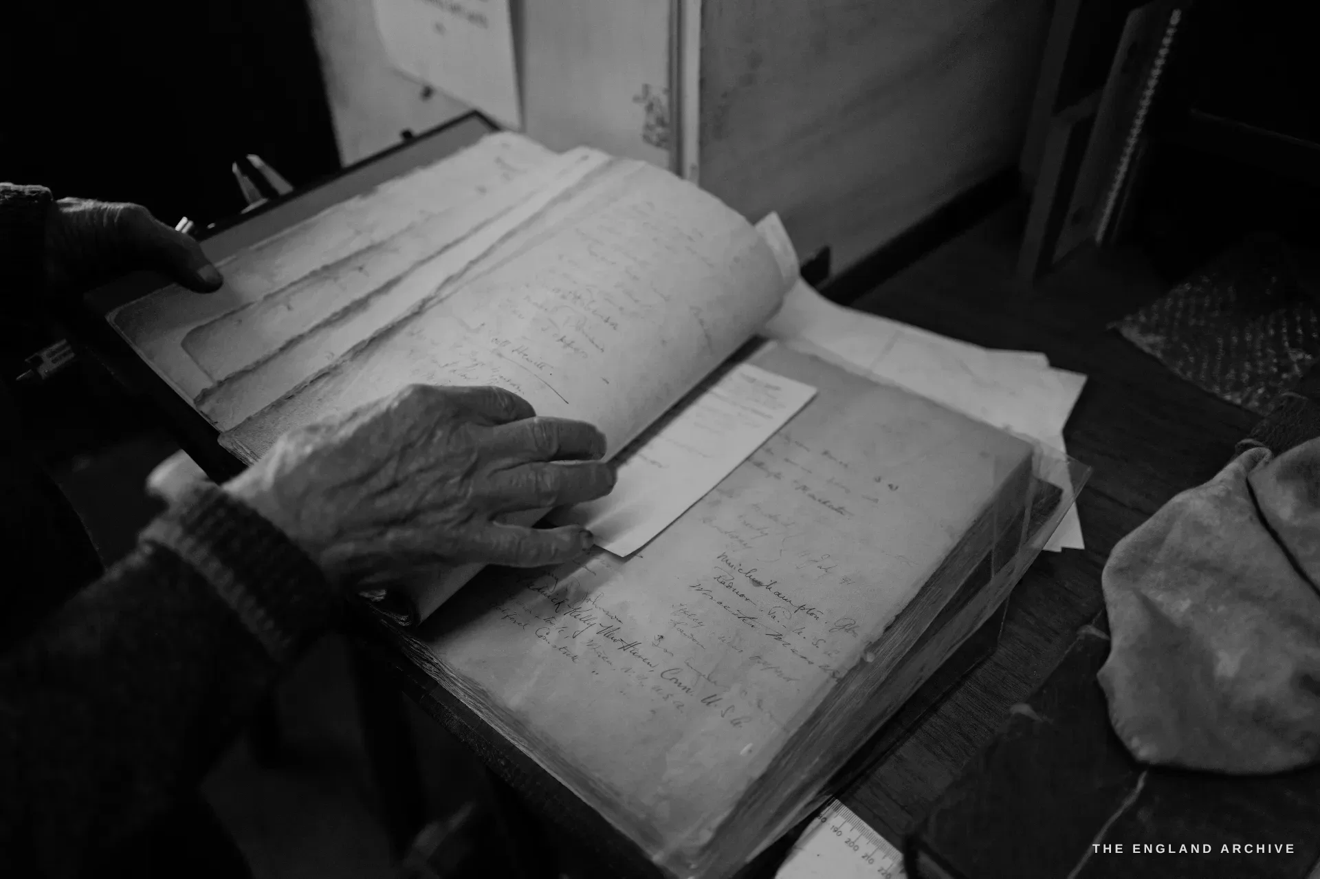 An elderly hand resting on and turning the leaves of a large handwritten order ledger, the pages dense with cursive entries and place-names.