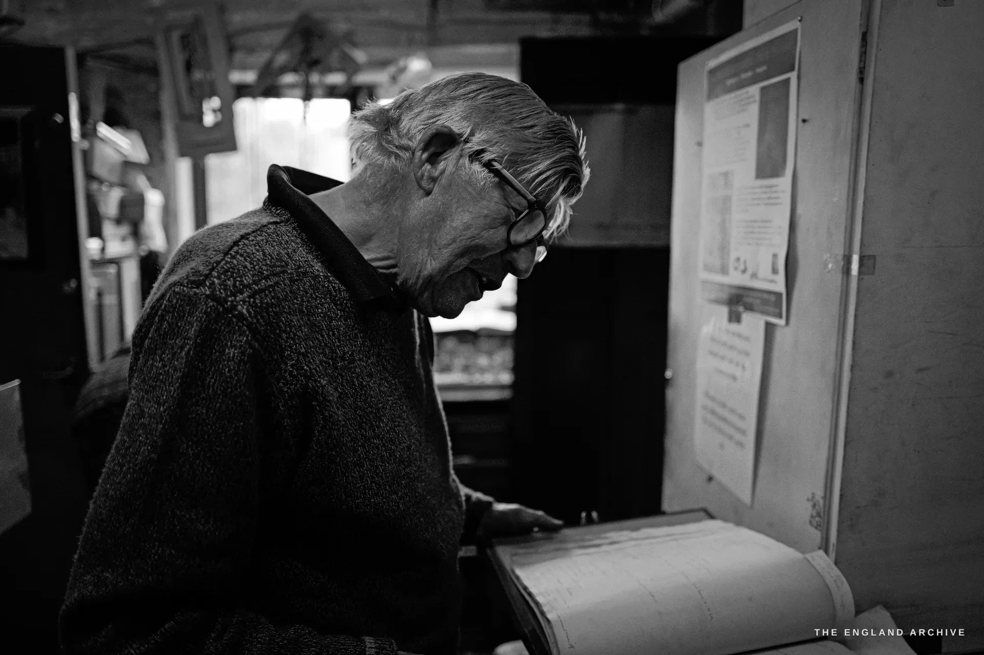 The elderly silversmith in profile reading over the open ledger at a stand, typed notices pinned to the cupboard door beside him.