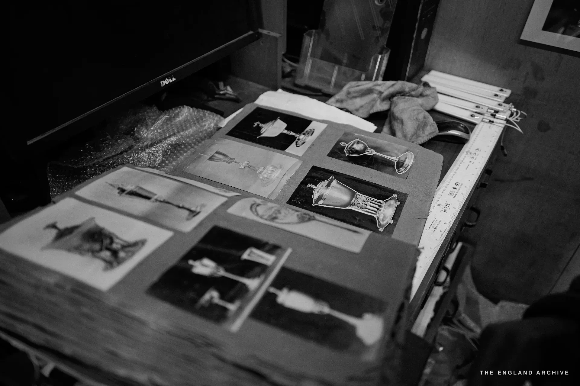 An open archive album of old monochrome catalogue photographs of Hart silverware - chalices, ewers and standing cups - laid in a grid on a cluttered desk.