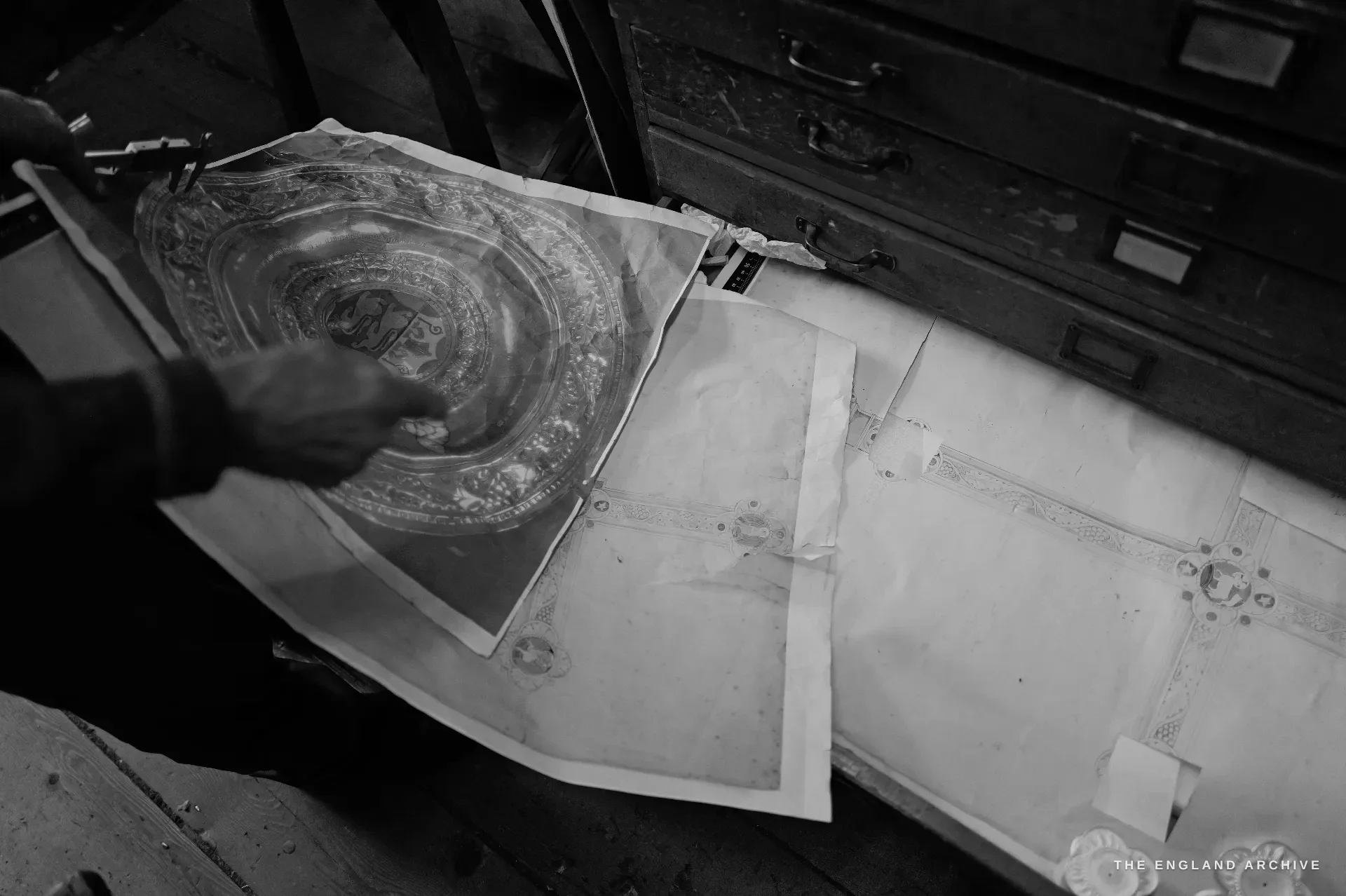 A hand lifting a large pencil drawing of a circular embossed dish over a plan-chest drawer of measured working drawings.