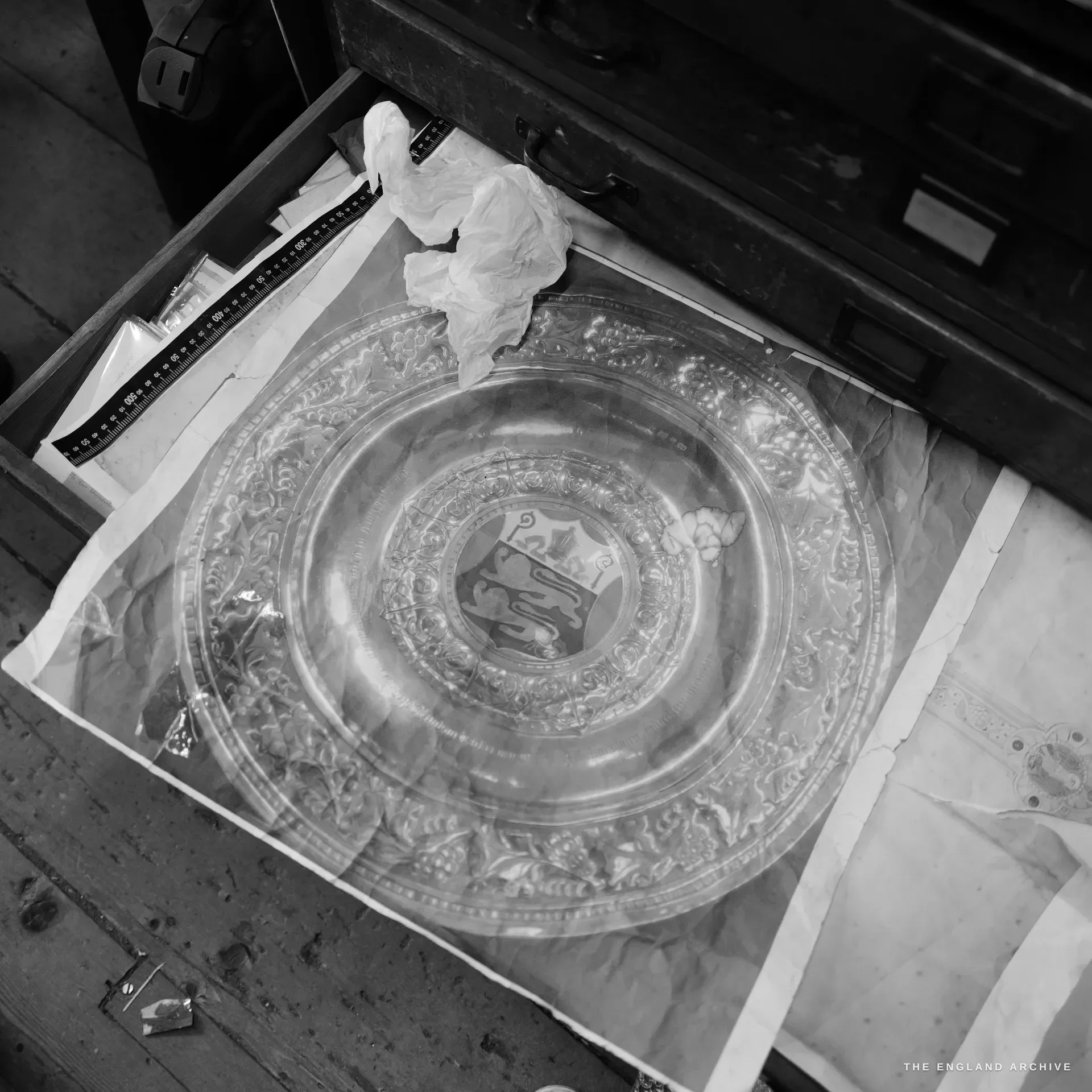 Seen from above, a large ornate silver charger lying in tissue in a drawer, a deep vine-and-grape repousse border encircling a central armorial shield.