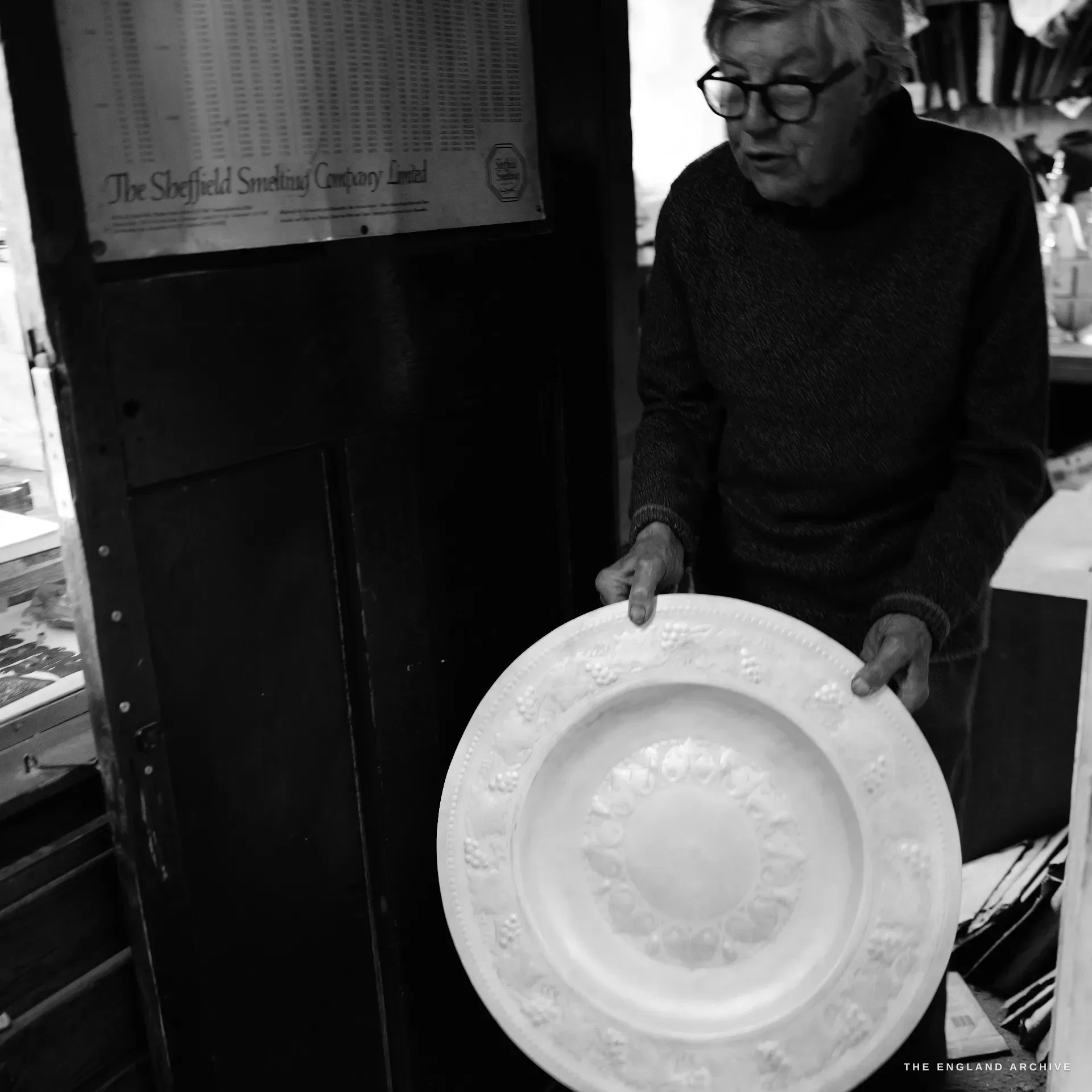 The elderly silversmith holds up a large embossed charger to show the camera, standing beside a tall cupboard bearing a printed Sheffield Smelting Company trade placard.