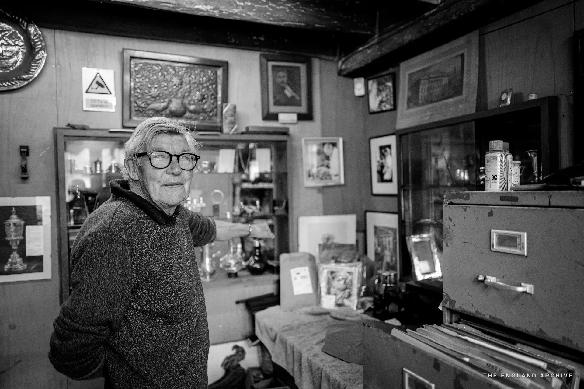 A three-quarter portrait of David Hart turning to face the camera in the workshop showroom, a wall of framed photographs and a circular Guild plaque behind him.