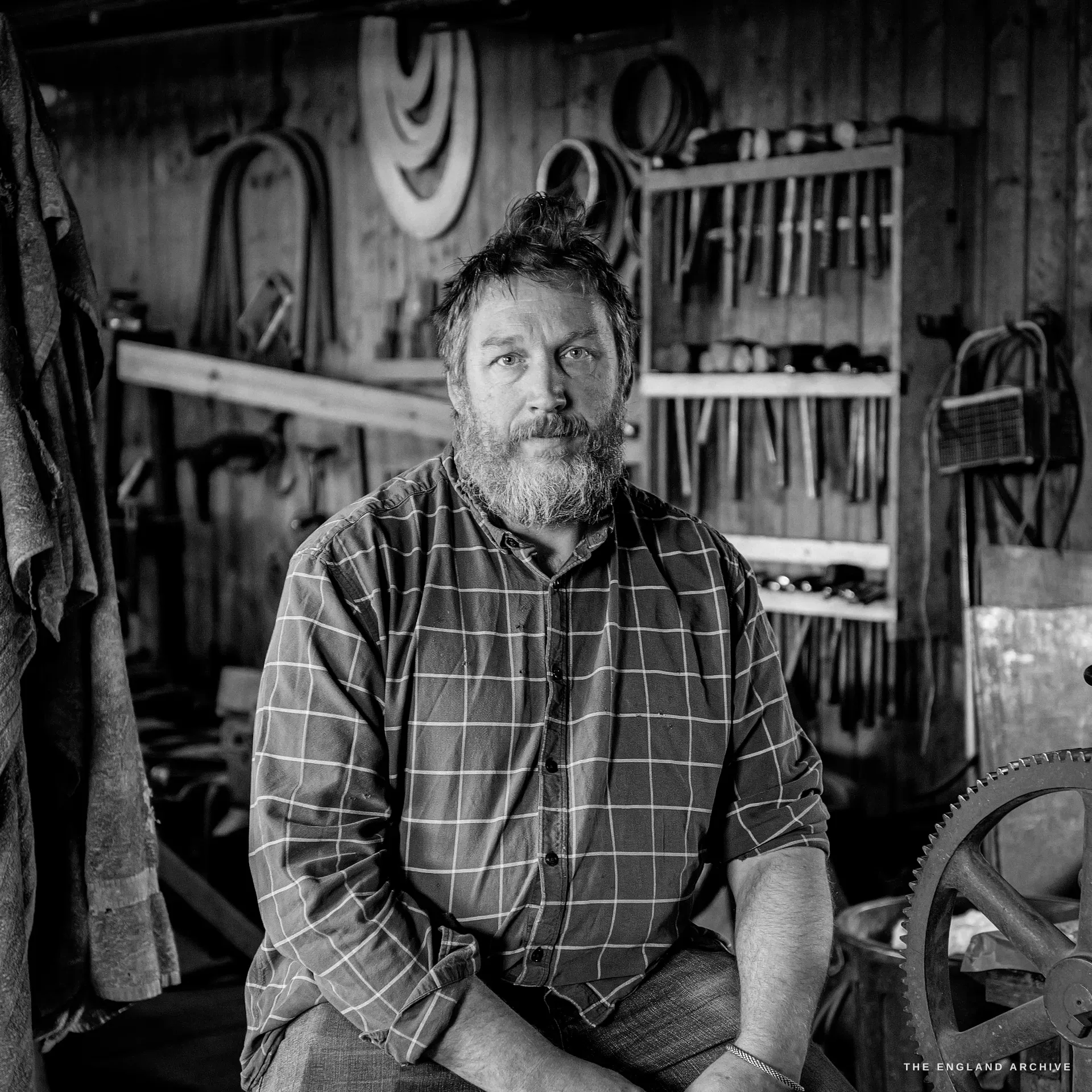 A square environmental portrait of Julian Hart, bearded with his hair in a top-knot, in a checked shirt, seated and facing the camera before the workshop's wall of hanging tools.
