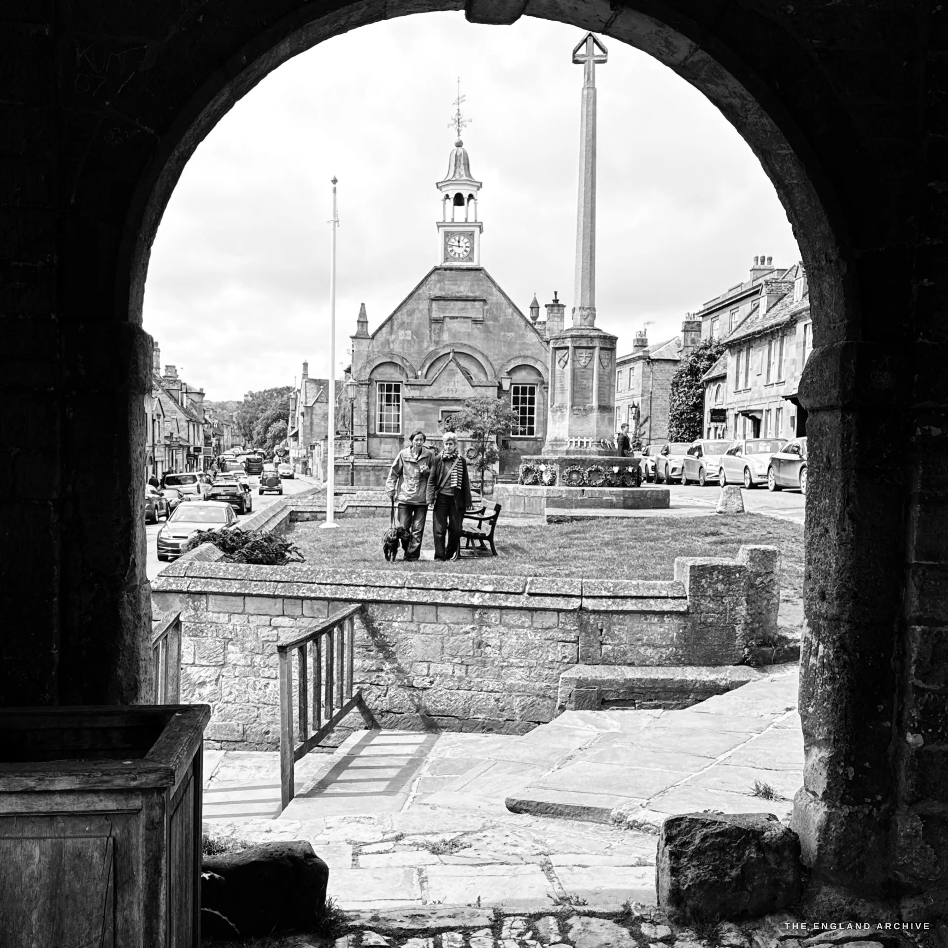 The green framed through a dark Market Hall arch - the gabled Market Hall, the war memorial cross and a flagpole beyond, with two people and a dog on the lawn and the High Street behind.