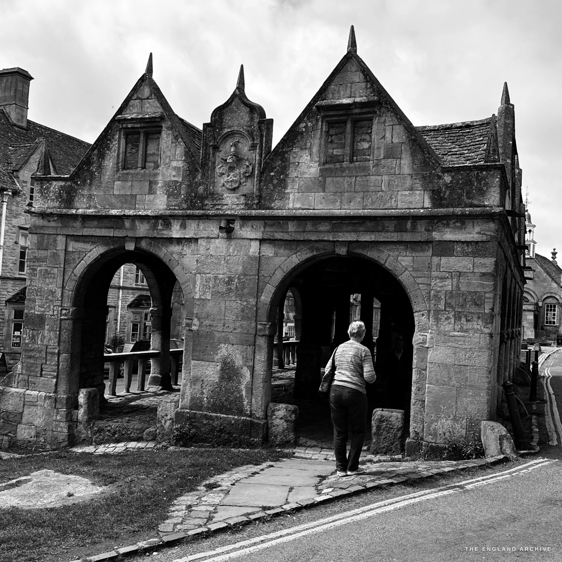 The 1627 open-arched Market Hall seen from the road, its gabled stone bays and pointed arches in side light, a woman walking into the right-hand arch.