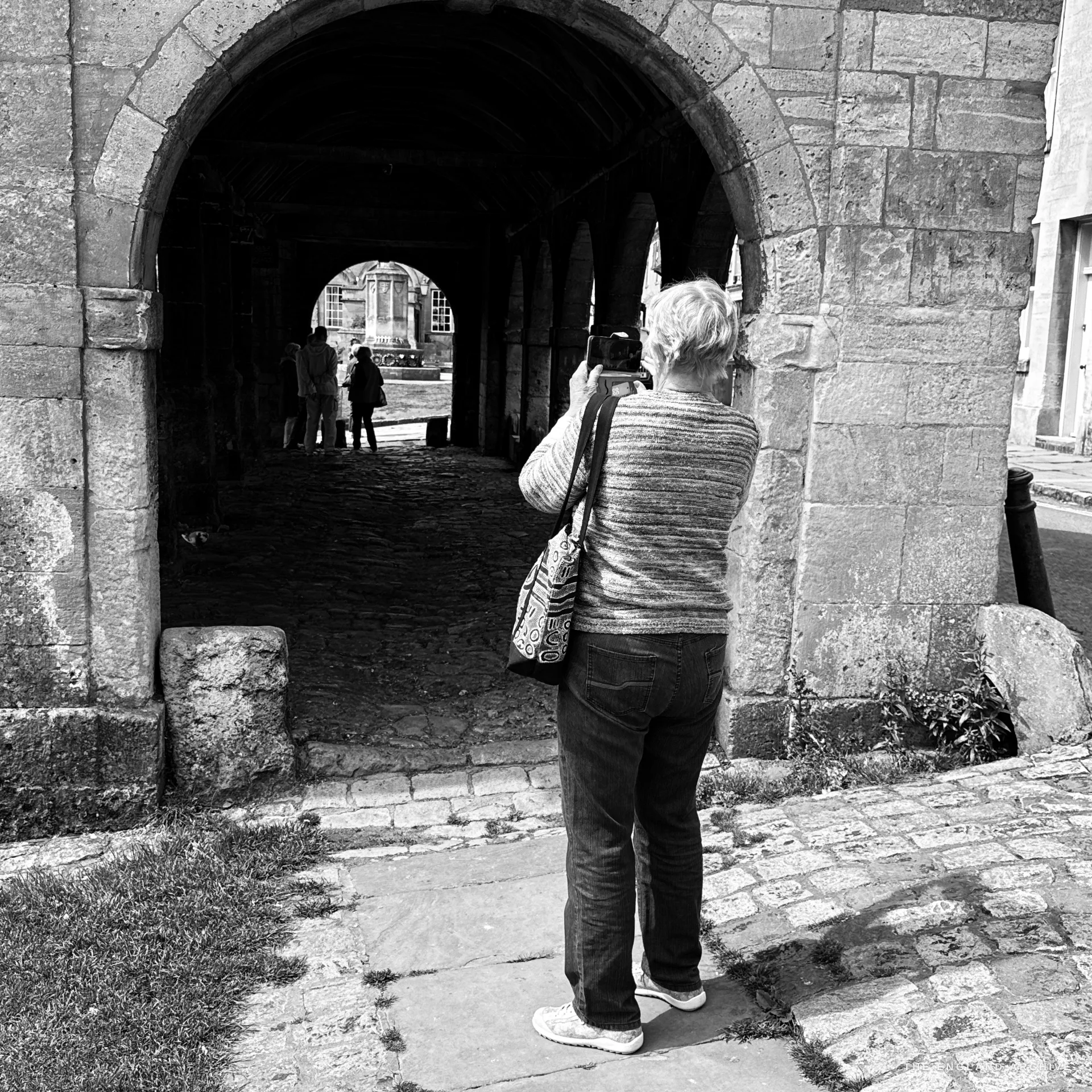 Inside the Market Hall, a silver-haired woman photographs through an arch toward the war memorial, silhouetted against the bright opening among the stone piers and cobbles.