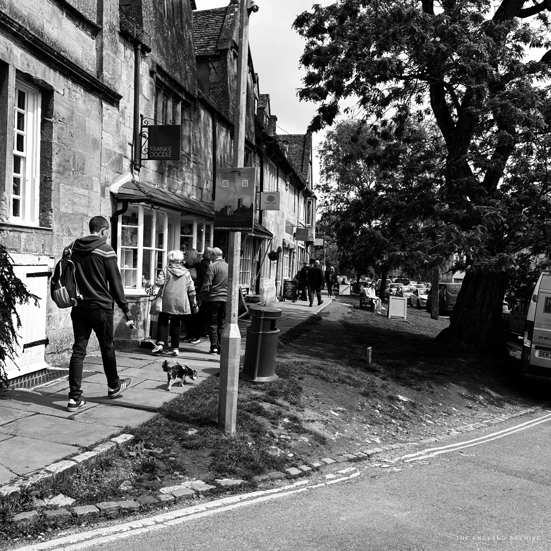 A High Street streetscape, a row of stone shops with a projecting sign, pedestrians and a small dog clustered at a shopfront, a large tree and parked van to the right.