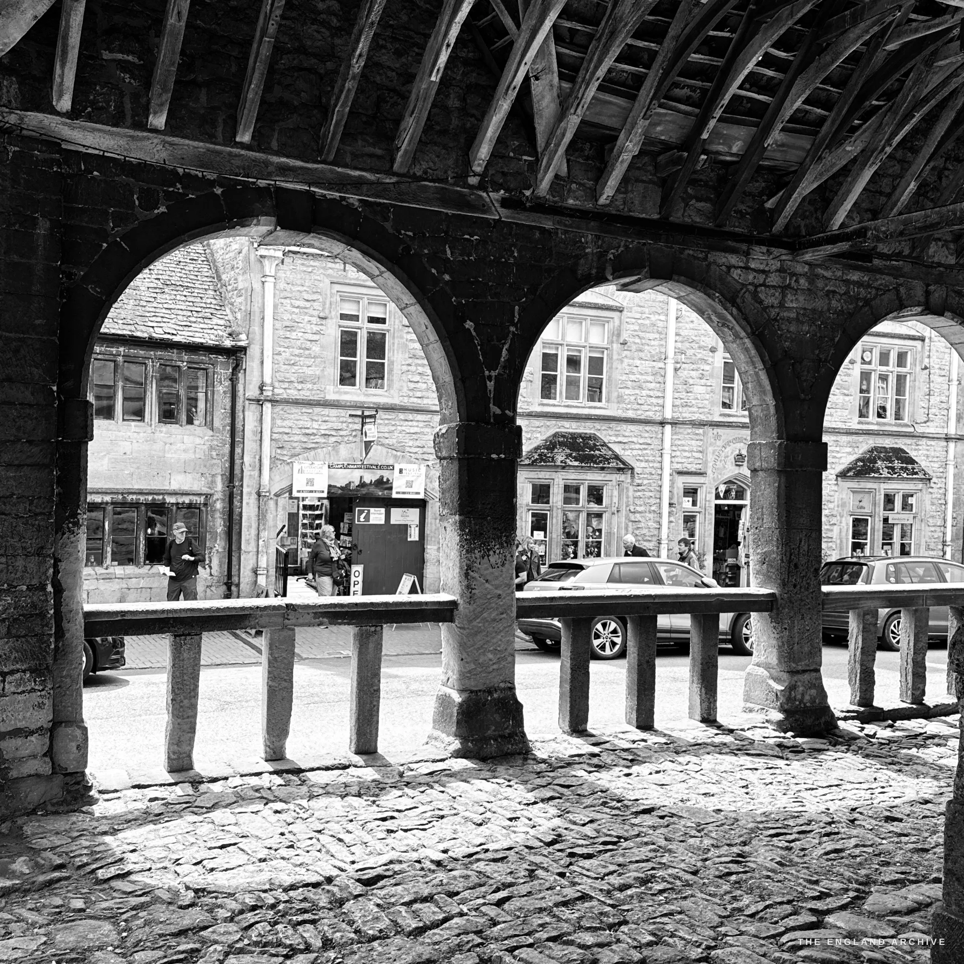 A view out from under the Market Hall's timber-raftered roof, through three stone arches across the cobbled floor to the shops and parked cars of the High Street opposite.