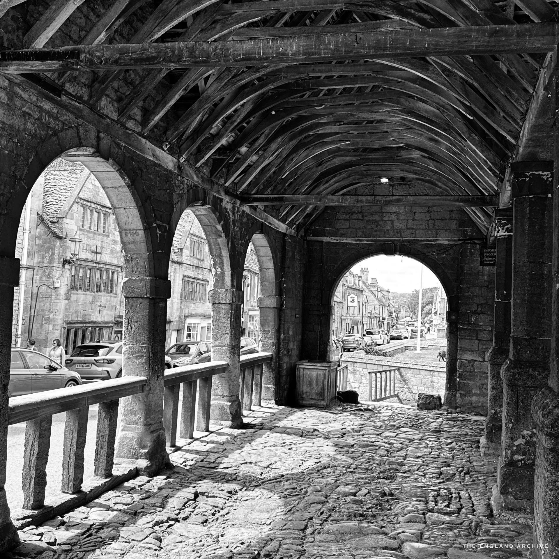 Inside the Market Hall, looking along its cobbled bay under the medieval timber wagon roof, arches opening to the street and a bright arch ahead framing the town beyond.