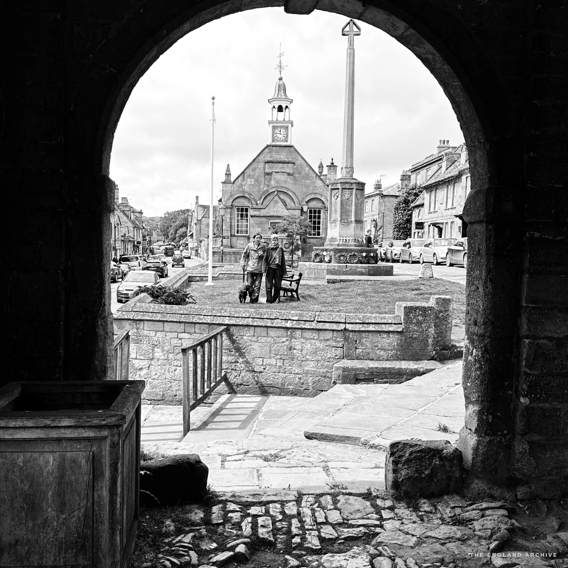 The Market Hall, war memorial cross and flagpole on the green framed through a Market Hall arch, two people with a dog posing on the lawn.