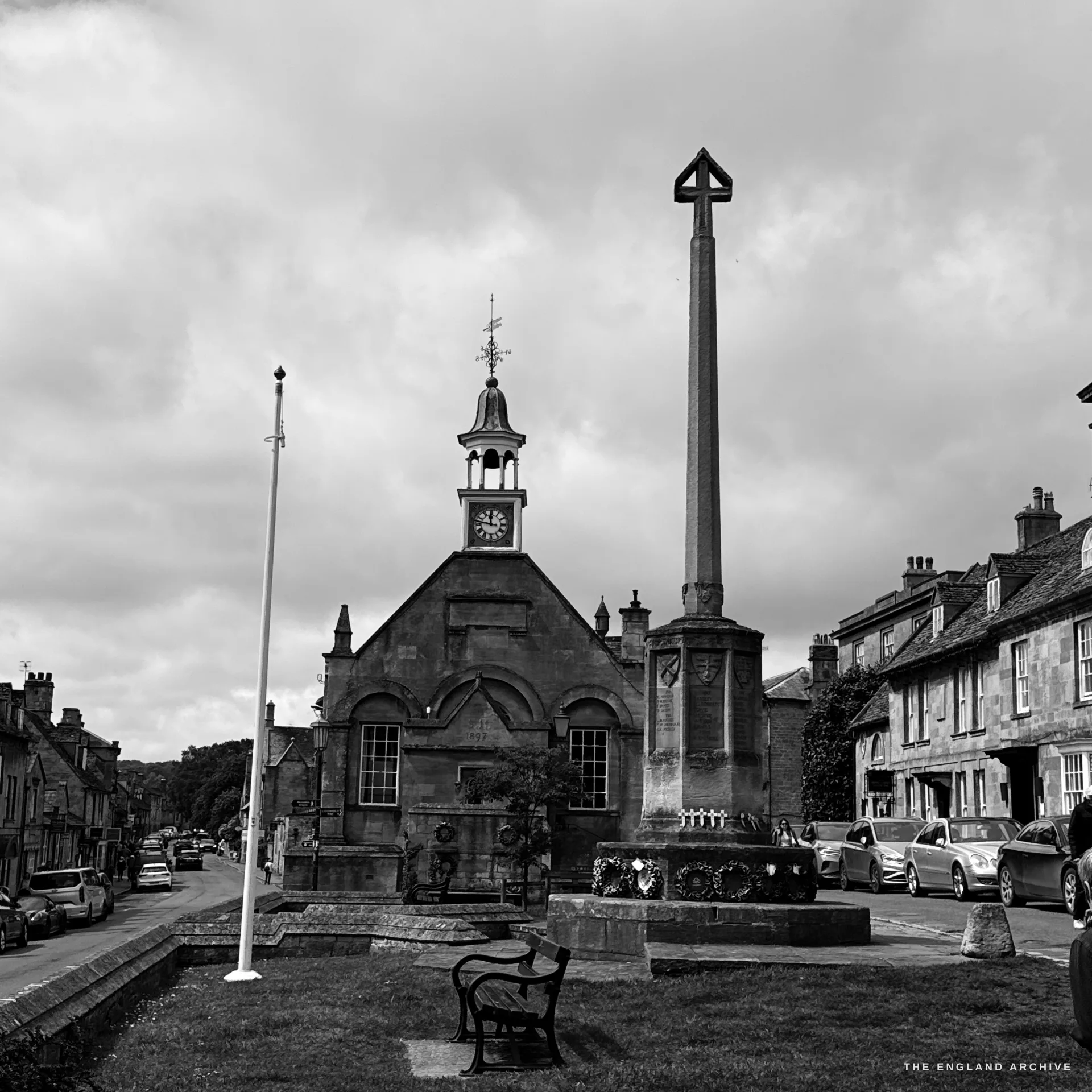 A wide townscape of the green: the gabled Market Hall with its clock-and-cupola turret on the left and the tall war memorial cross on the right, a bench on the grass and the stone High Street receding under a heavy sky.