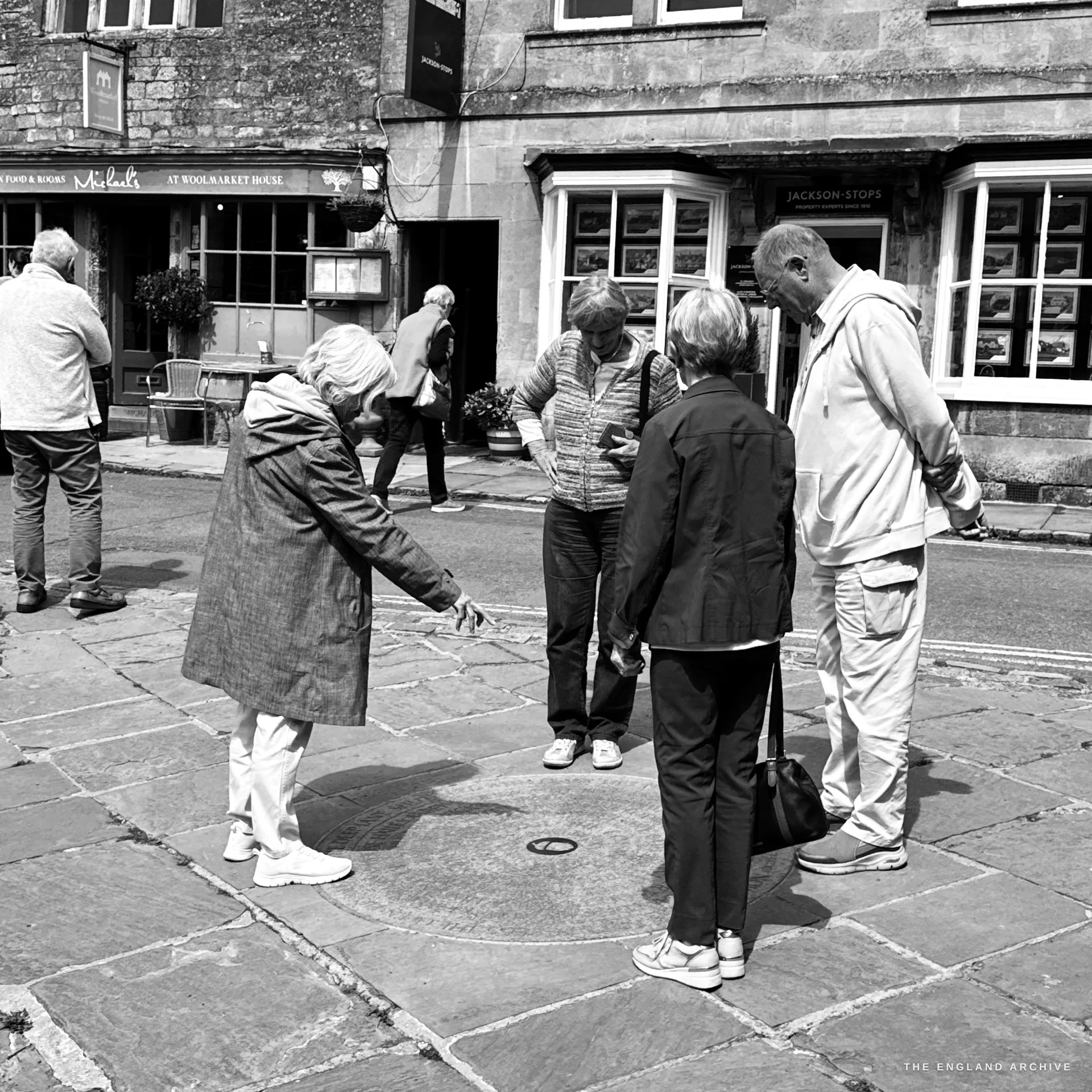 A group of older people on the wide flagstone pavement examining a circular bronze pavement marker set in the stone, the Jackson-Stops office and a pub behind them.