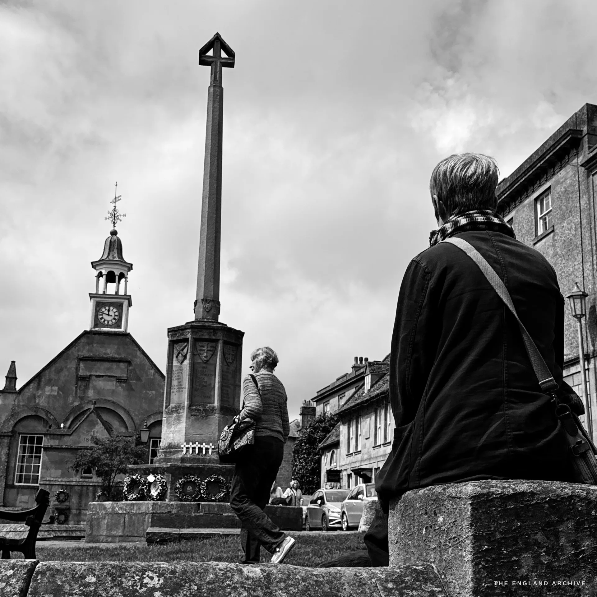 A low-angle view of the war memorial cross, a person seated on the stone plinth in the foreground and another reading the memorial, the Market Hall cupola to the left.