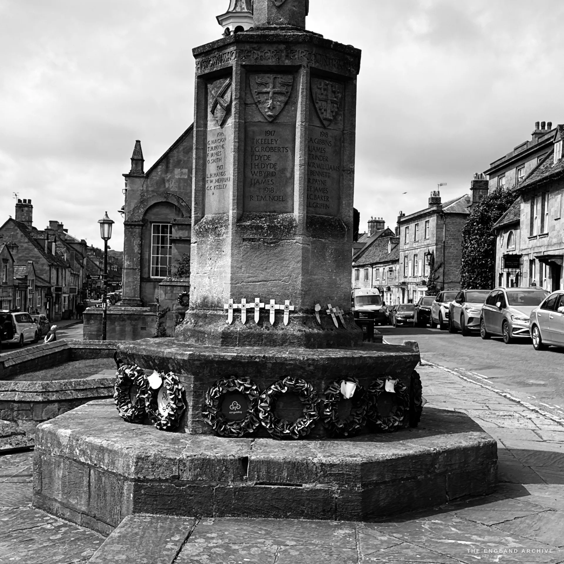 A close study of the octagonal war memorial base, its inscribed name panels and carved heraldic shields above, a row of small white remembrance crosses and laid poppy wreaths around the foot.