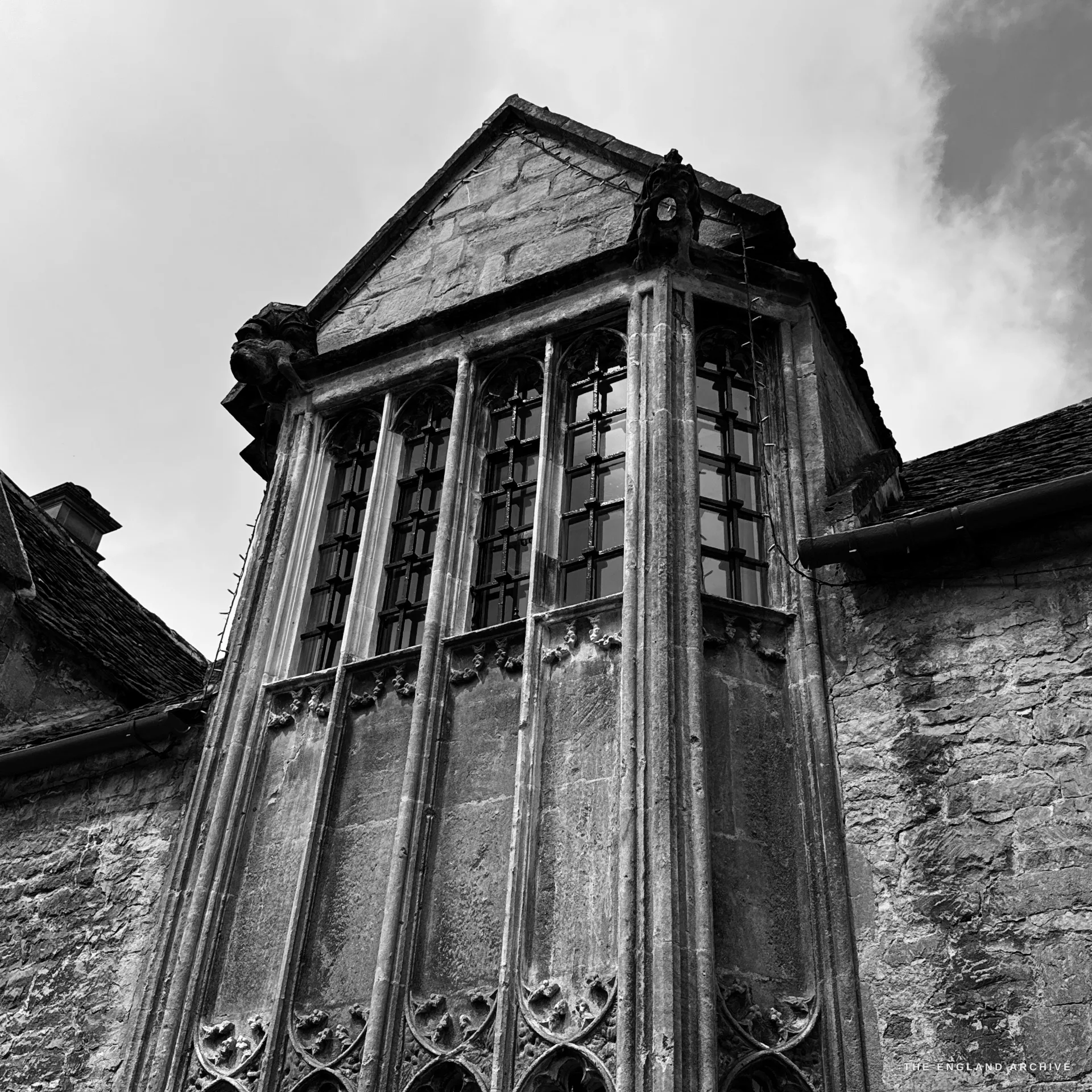 An upward view of a tall mullioned and transomed gabled stone oriel window in the manner of Grevel's House, a carved gargoyle to the left, set against the sky.