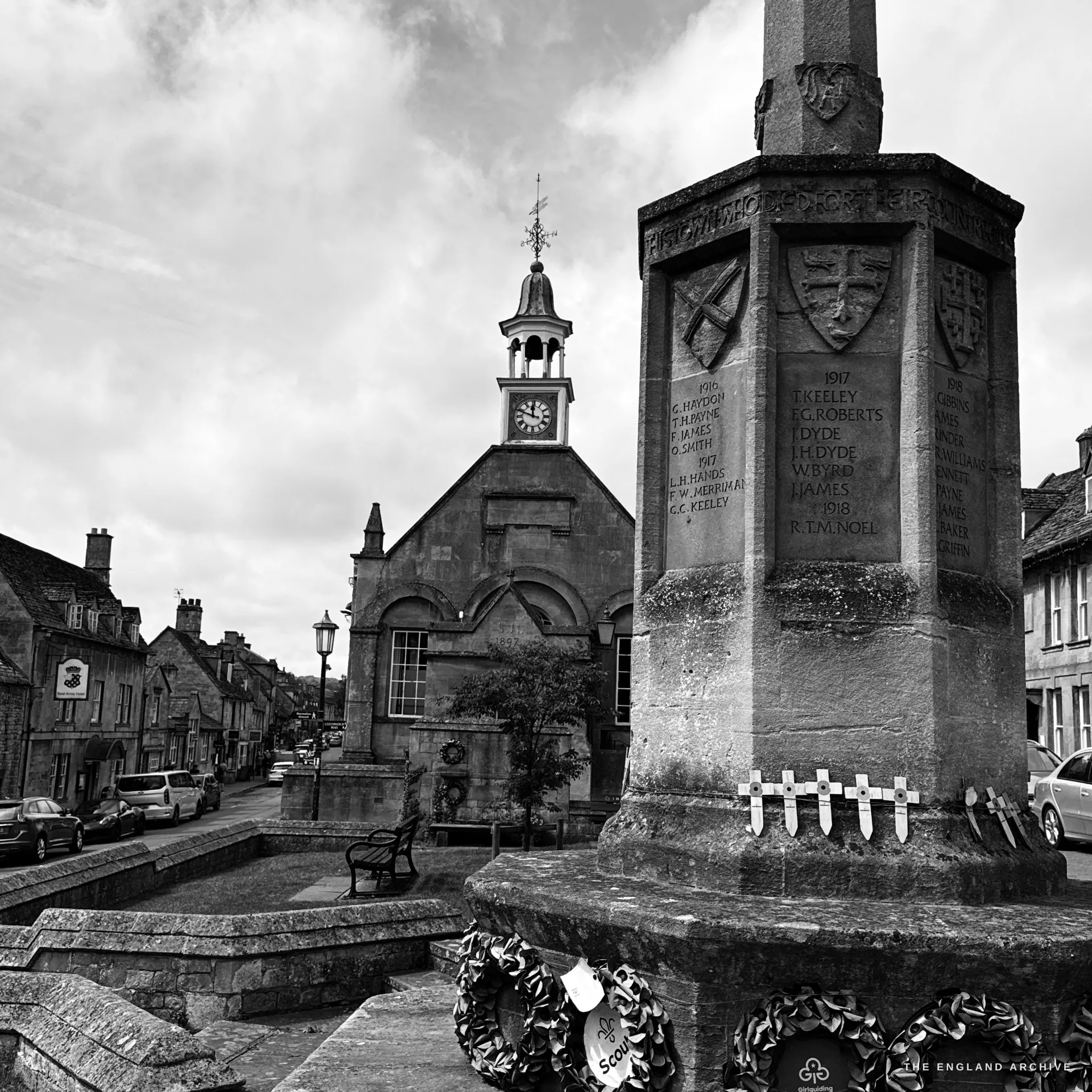 The war memorial cross close at the right with its heraldic shields and name panels, the Market Hall cupola, a bench and the descending stone High Street to the left under a grey sky.