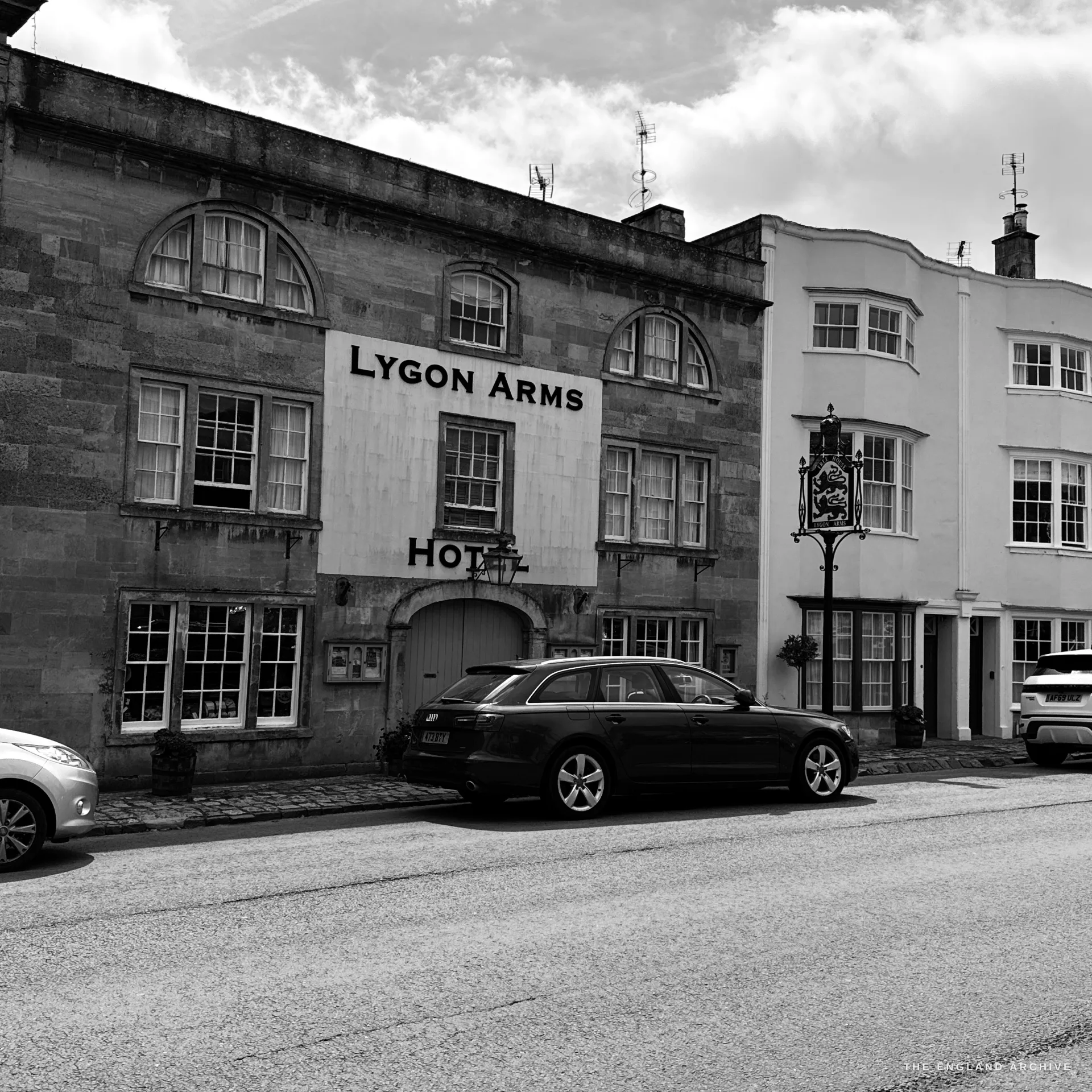 The honey-stone frontage of the Lygon Arms Hotel with rounded dormer windows and a hanging heraldic sign bracket, a car parked on the road in front.