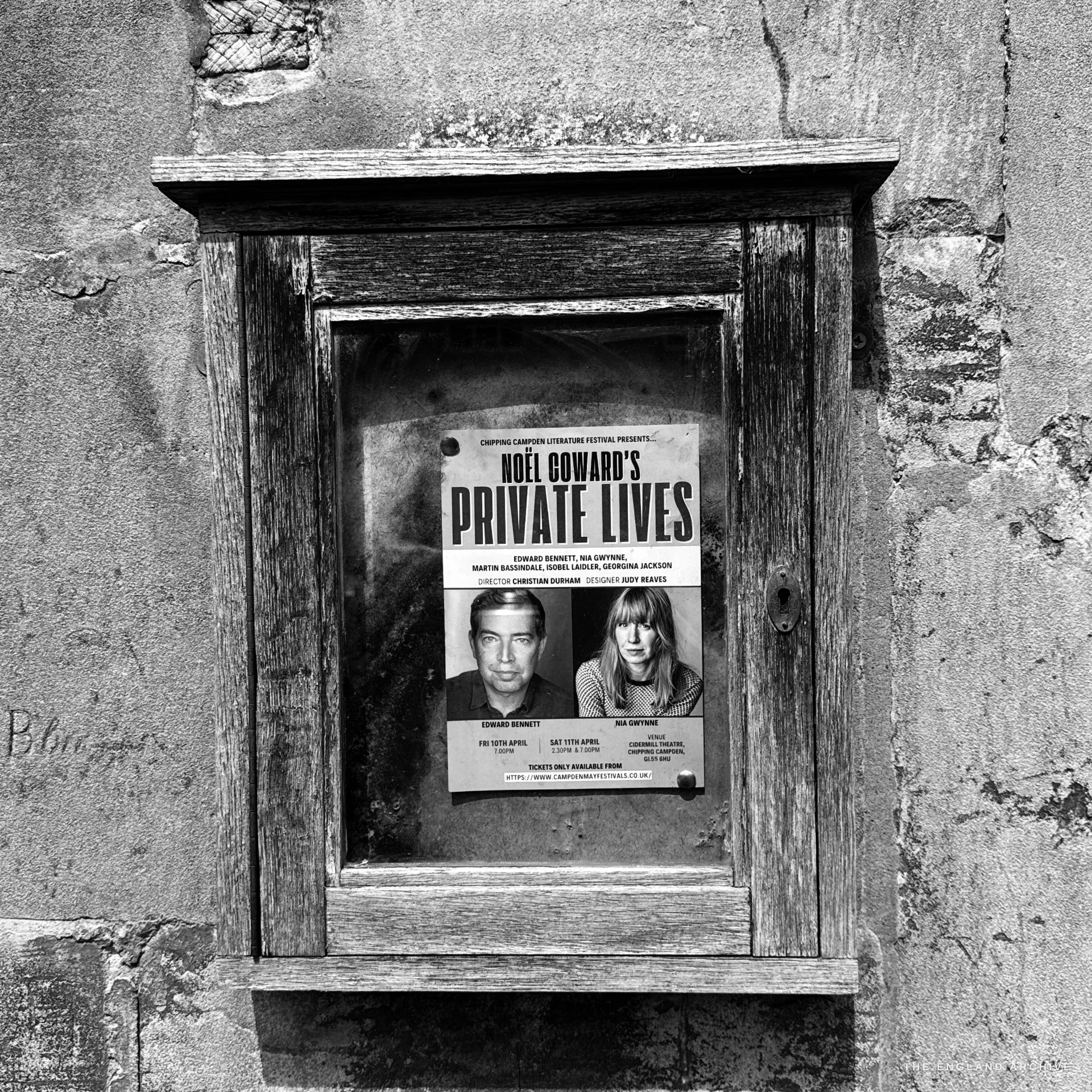 A weathered timber glazed notice case on lichened stone holding a printed poster for the Chipping Campden Literature Festival.