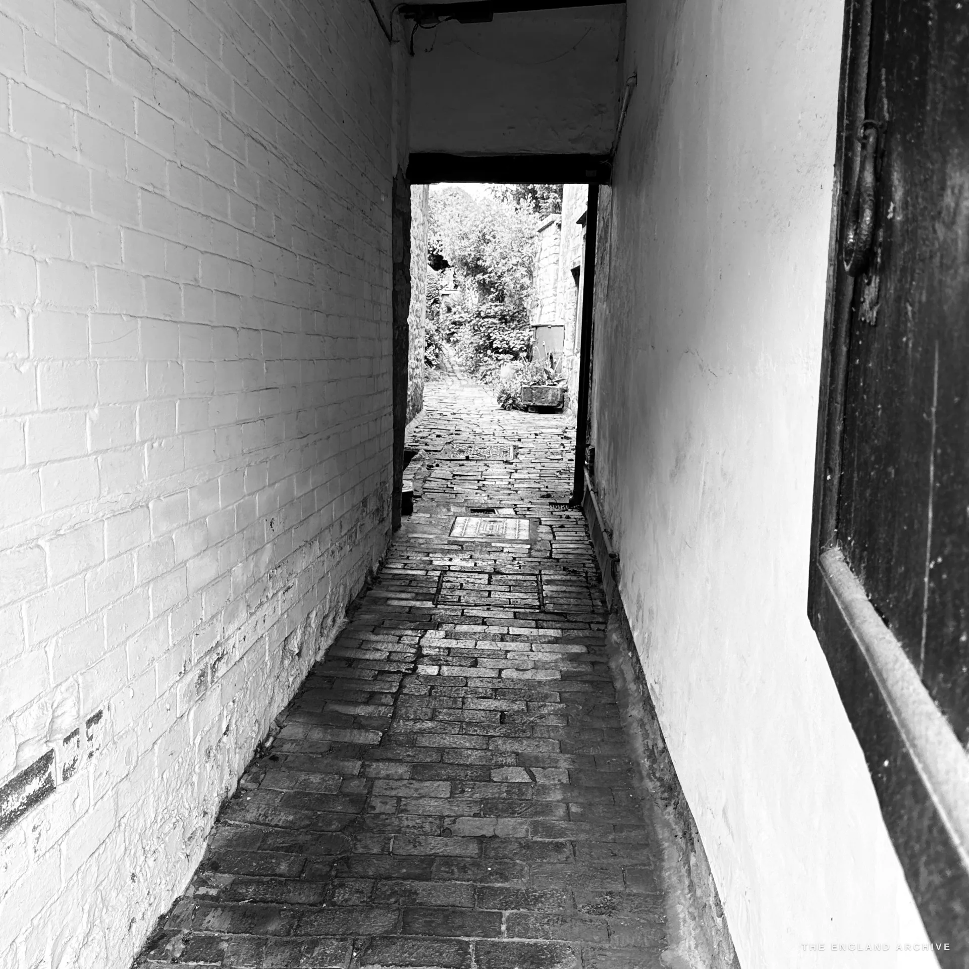 A narrow whitewashed pedestrian alley with a worn brick-paved floor leading toward a sunlit garden and greenery at its far end.