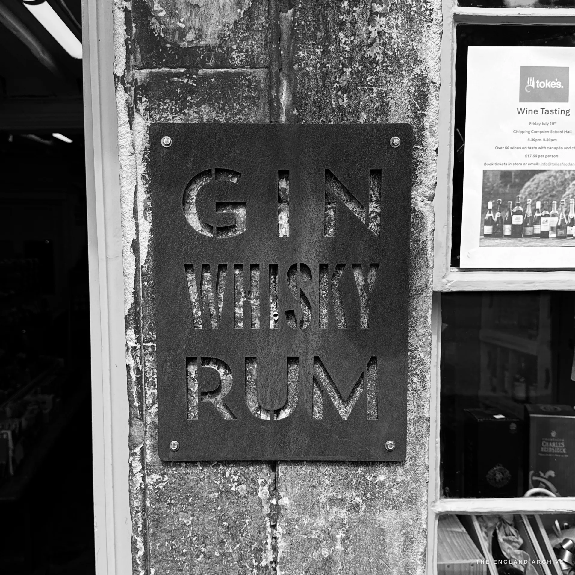 A laser-cut steel shop sign reading Gin, Whisky, Rum fixed to a stone pier beside a wine merchant's window of bottles and a tasting notice.