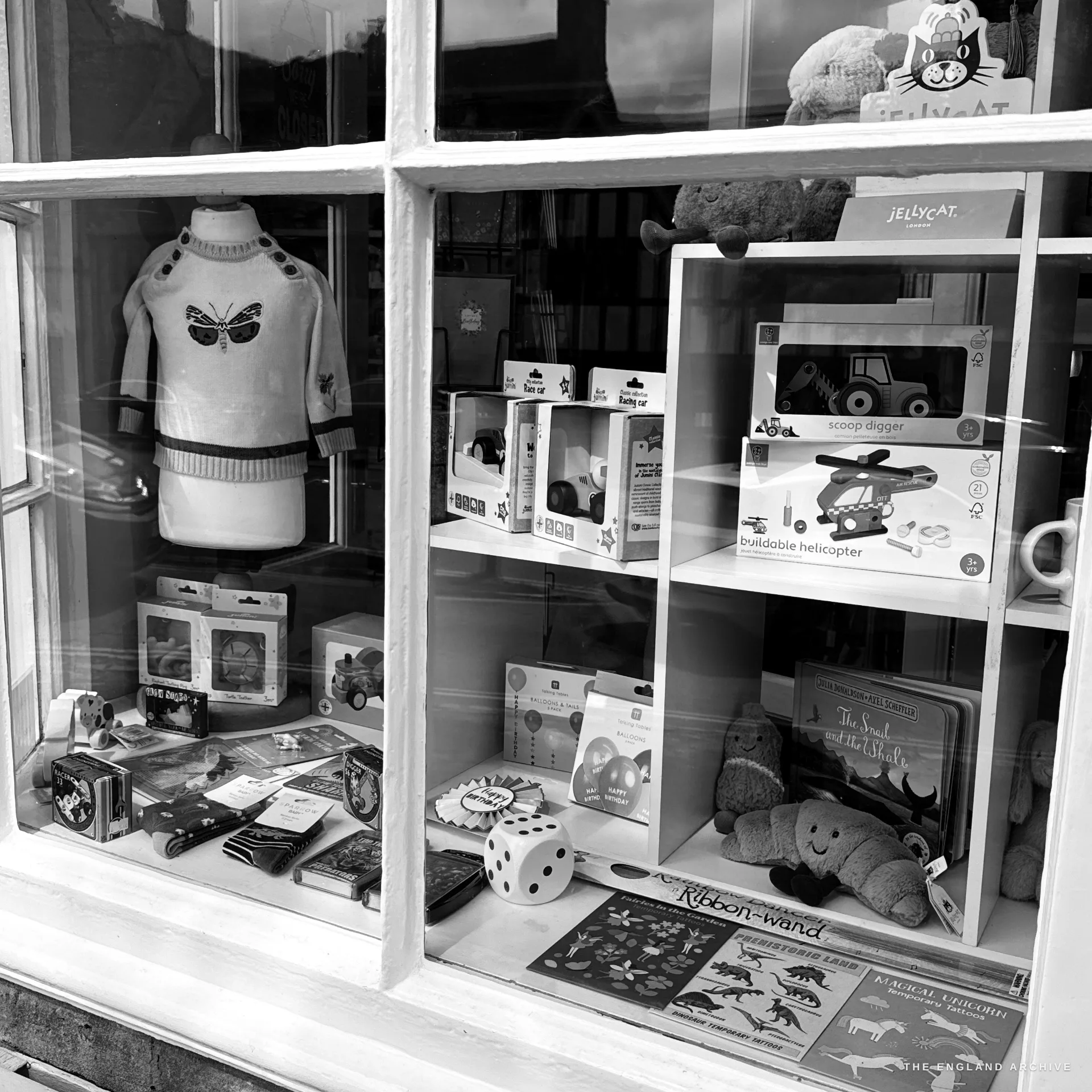 A toy and gift shop window display behind white glazing bars, with a child's jumper, boxed toys, soft toys and books.