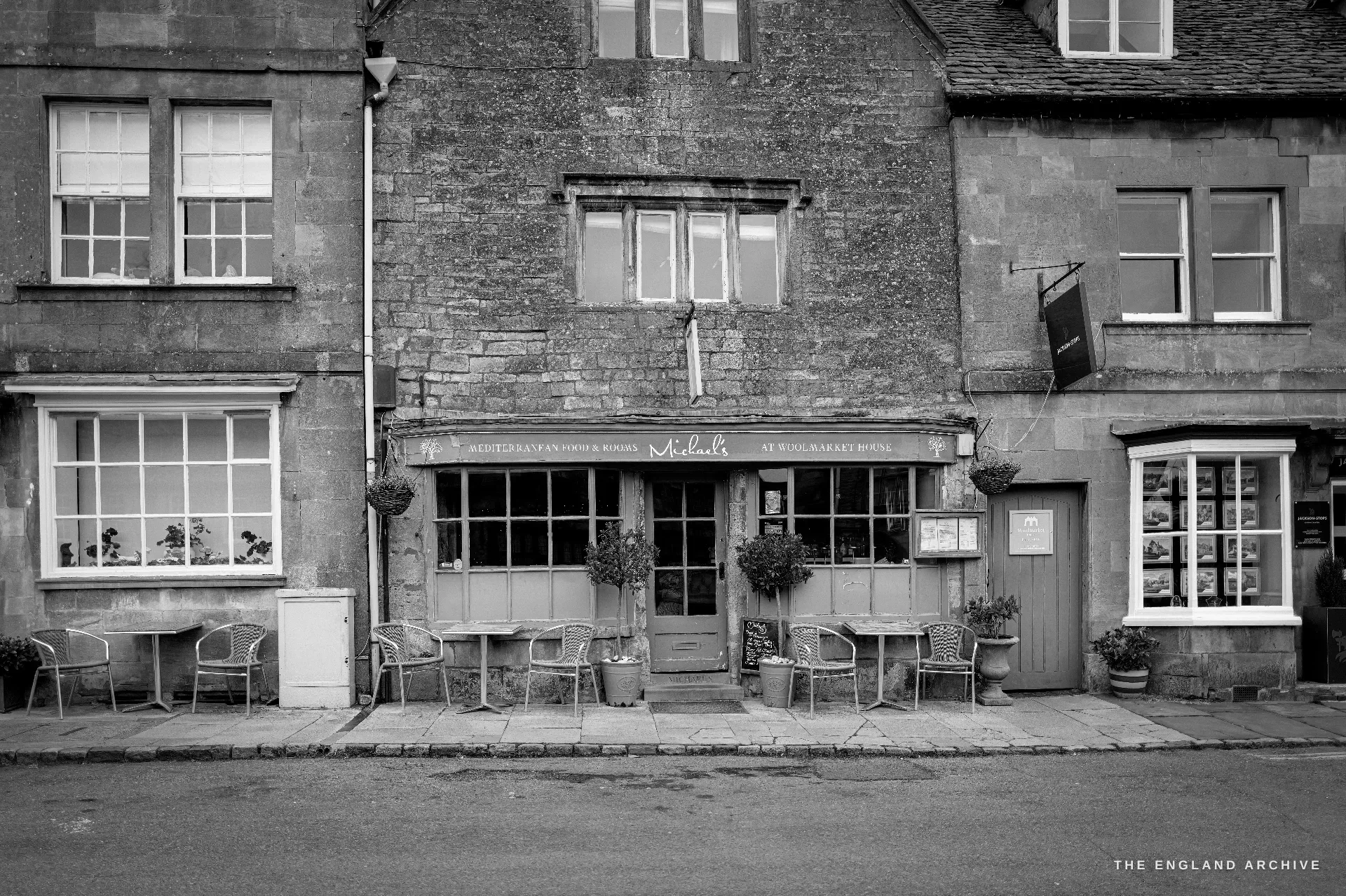 A stone-and-brick cafe front with bay windows, outdoor tables and chairs and hanging baskets, its painted fascia sign above the door.