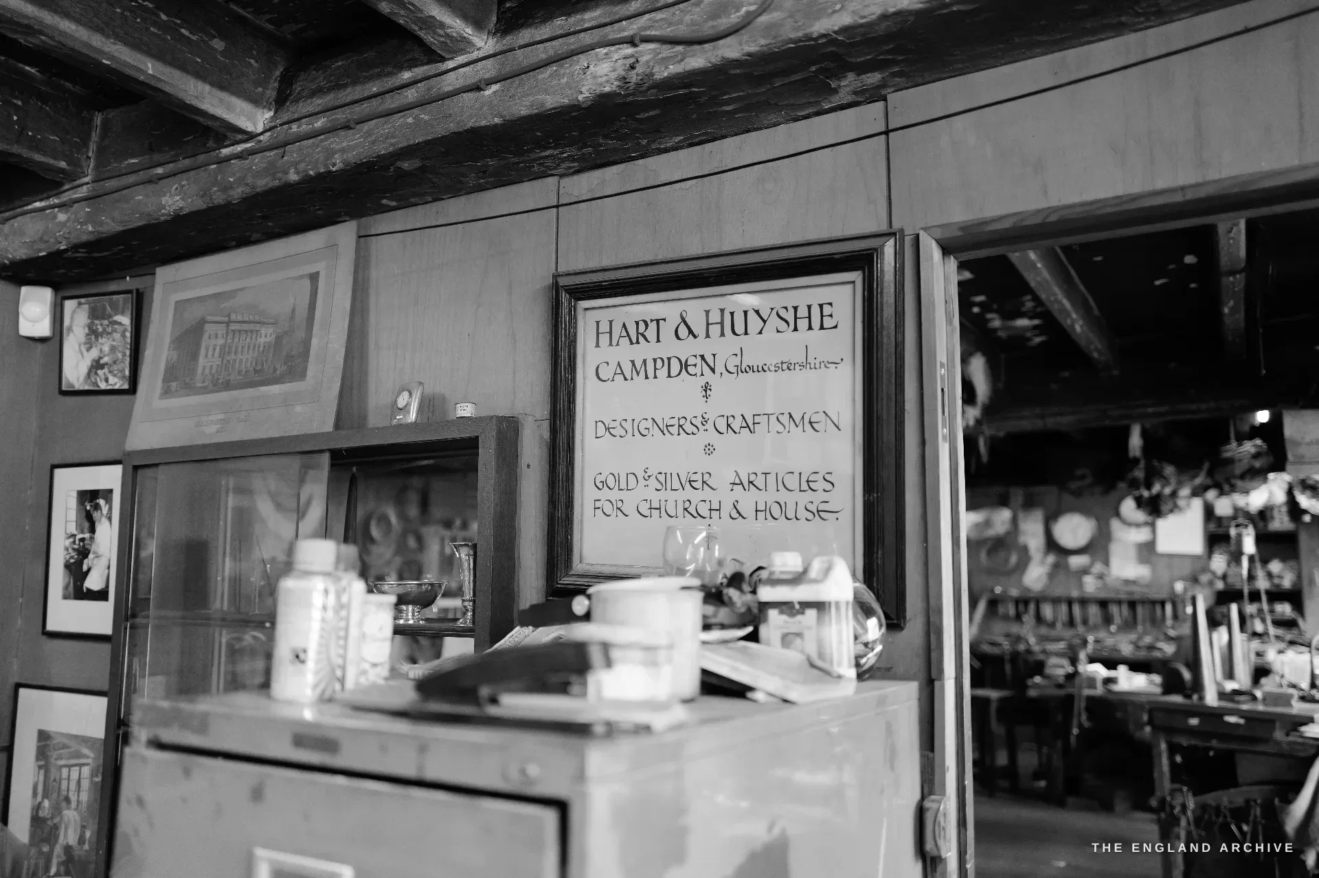 A hand-lettered framed trade sign hung high in the showroom reading Hart and Huyshe, Campden, Designers and Craftsmen, Gold and Silver Articles for Church and House.