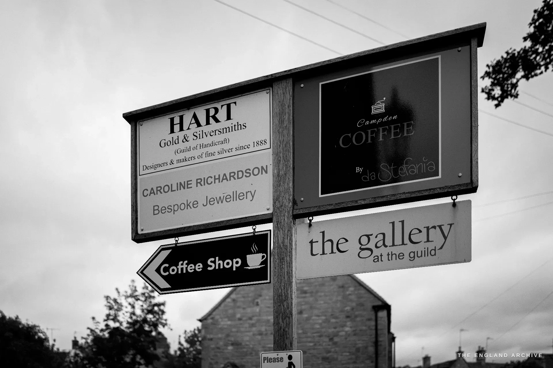 A cluster of hanging painted trade signs on a timber post by the road - the Hart workshop sign, a coffee-shop sign and a gallery sign - against an overcast Cotswold sky.