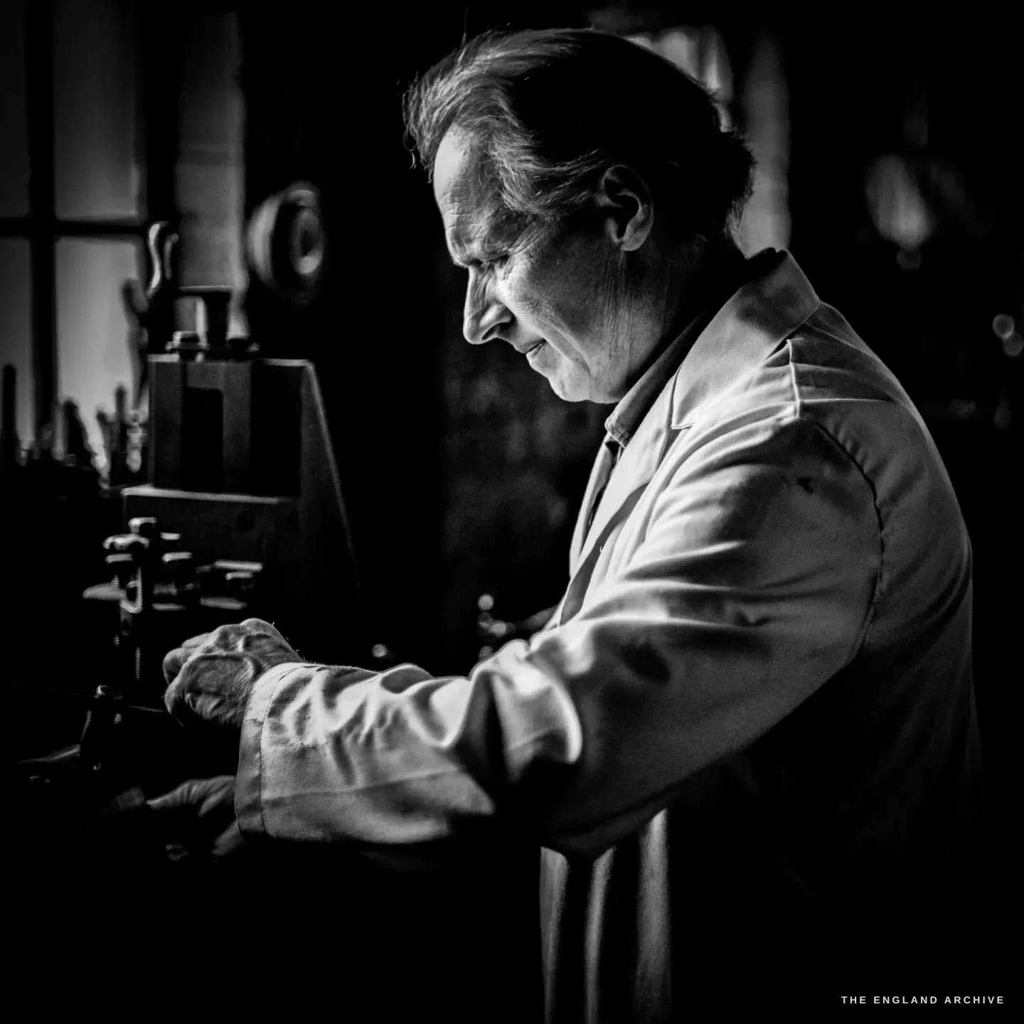Haydn Hill working at a machine by a window, both hands on the work, side-lit against the daylight, the dark workshop around him.